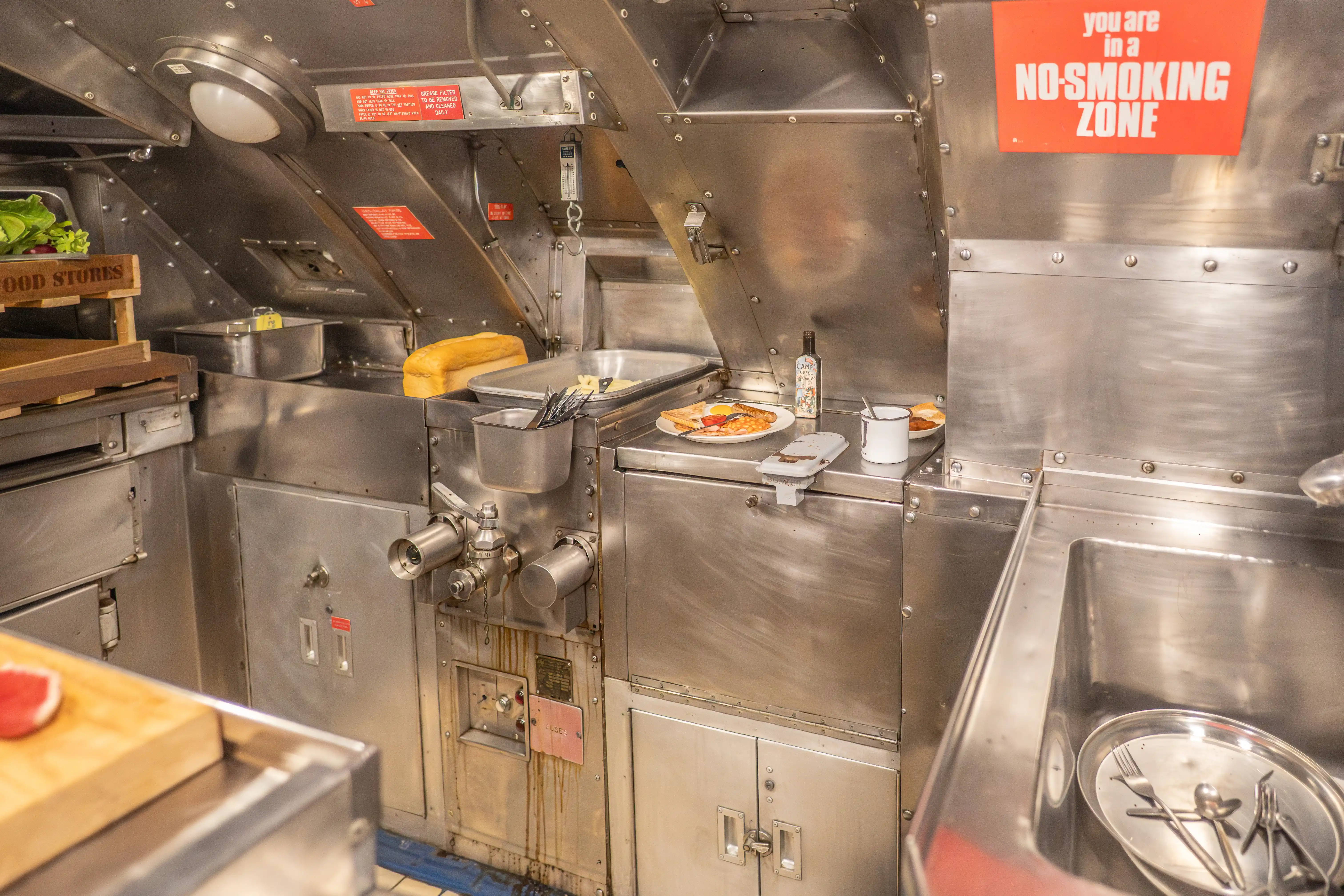 The image depicts a compact, metallic kitchen area, likely within a confined space. The kitchen is equipped with various cooking utensils and food items, including a bottle, a plate with food, and a container of utensils. A prominent sign indicates that it is a no-smoking zone. The setup includes storage areas and preparation surfaces, emphasizing functionality in a limited space.