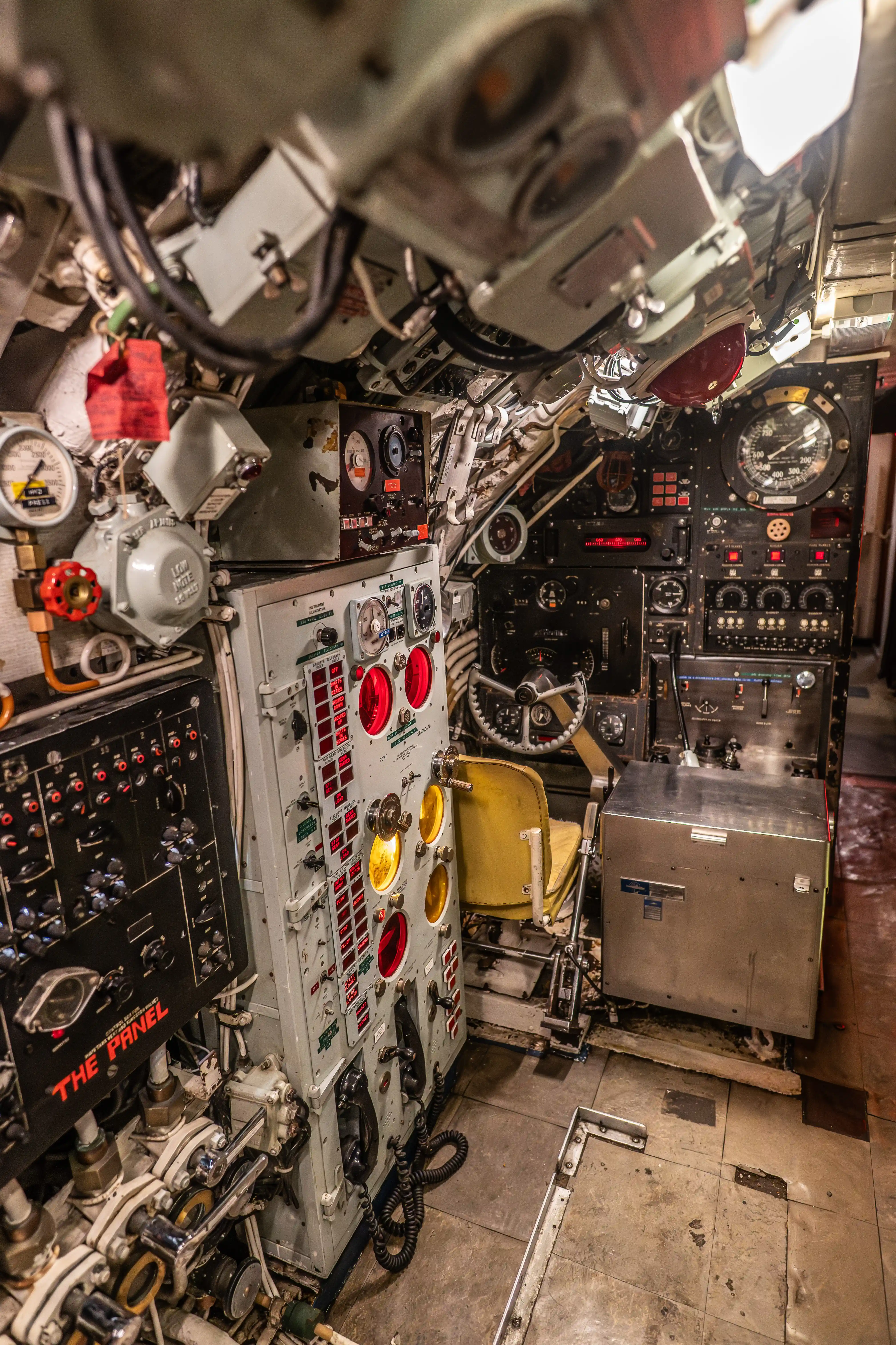 The image depicts the interior of a control room, likely from a submarine or a similar vessel. It is filled with various control panels, gauges, dials, and equipment, including a yellow chair positioned in front of a central console. The space is compact and densely packed with machinery and instruments, suggesting a highly functional and technical environment. The presence of numerous labeled switches, buttons, and indicators highlights the complexity and operational nature of the room.