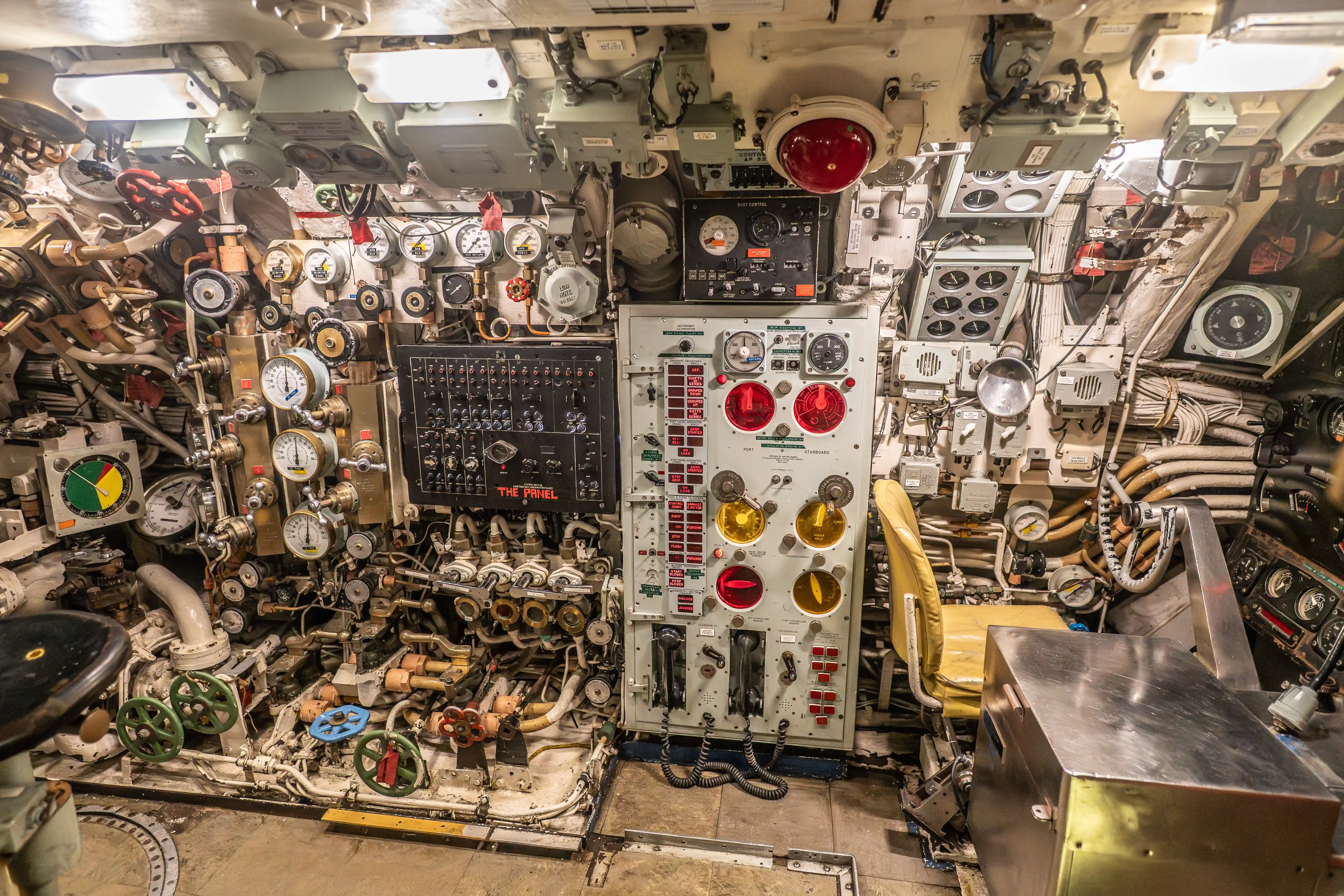 The image depicts the interior of a complex control room, likely from a submarine or a similar vessel. It is filled with numerous gauges, dials, valves, and control panels, indicating a highly technical and operational environment. The equipment appears to be well-used, suggesting it is either historical or actively in use. The space is tightly packed with various mechanical and electronic instruments, highlighting the intricate nature of managing such a vessel. A chair is visible, indicating a station for an operator or crew member.