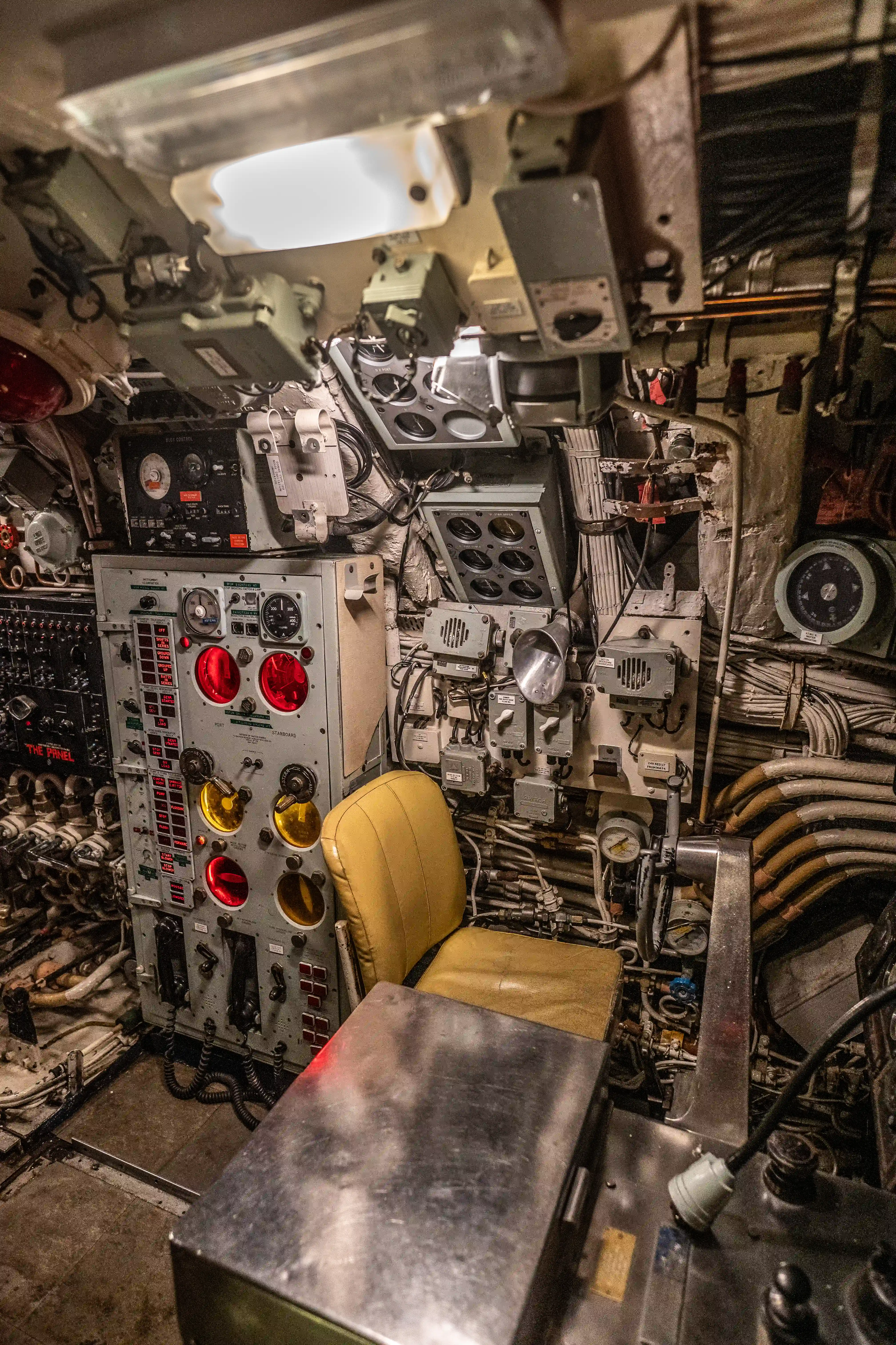 The image depicts the interior of a control room, likely from a submarine or similar vessel. The space is filled with various control panels, gauges, and equipment, including a prominent control console with multiple switches, buttons, and illuminated indicators. A yellow seat is positioned in front of the main control panel, and there are numerous pipes, wires, and mechanical components visible throughout the room. The environment appears to be compact and highly functional, designed for monitoring and operating complex systems.