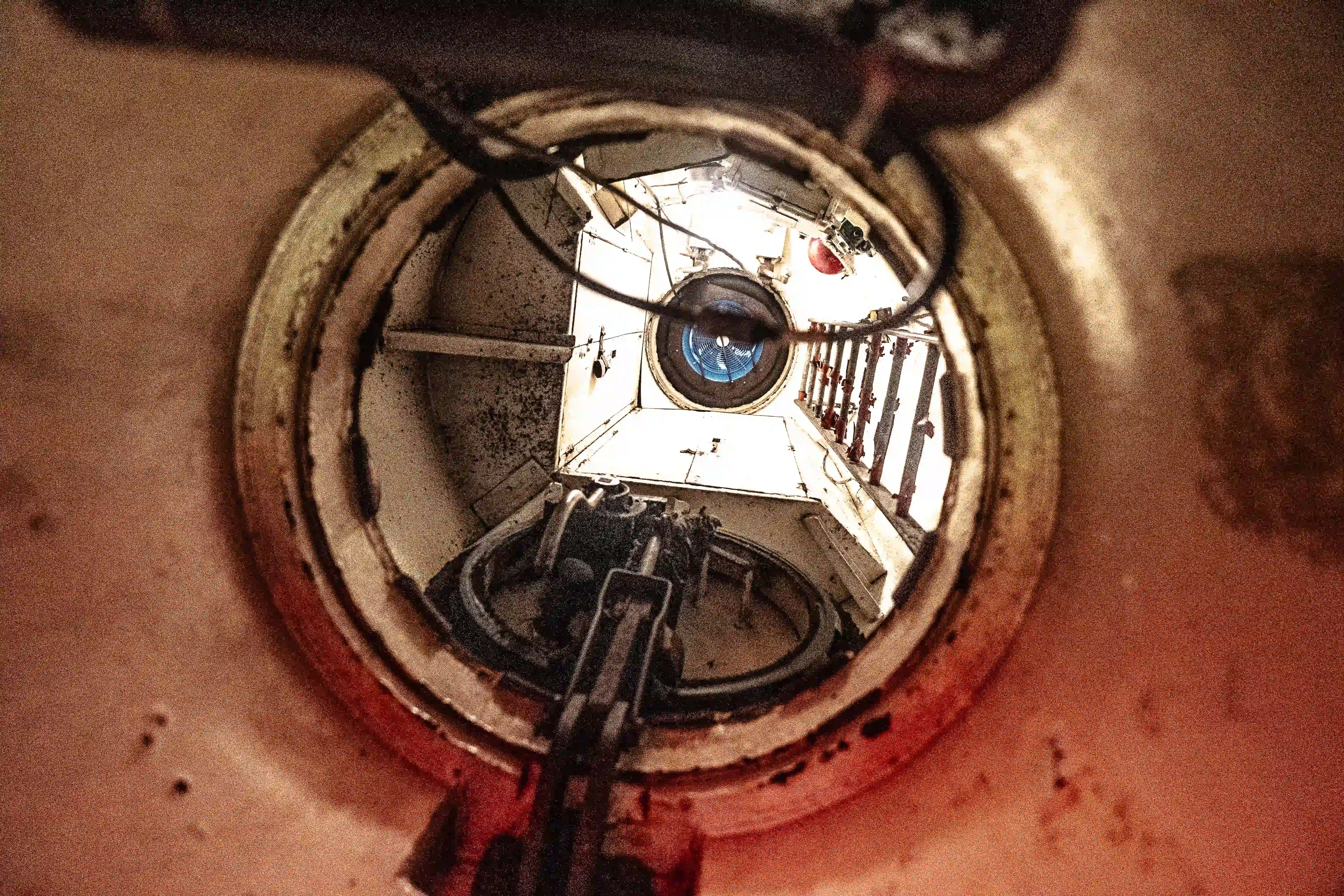 The image depicts an interior view of a cylindrical structure, possibly a submarine or industrial tank. The view is from within a hatch or opening, looking upwards towards another hatch or opening above. The surroundings are metallic and rusty, indicating age or exposure to the elements. Various mechanical components and railings are visible, suggesting a functional and industrial environment.