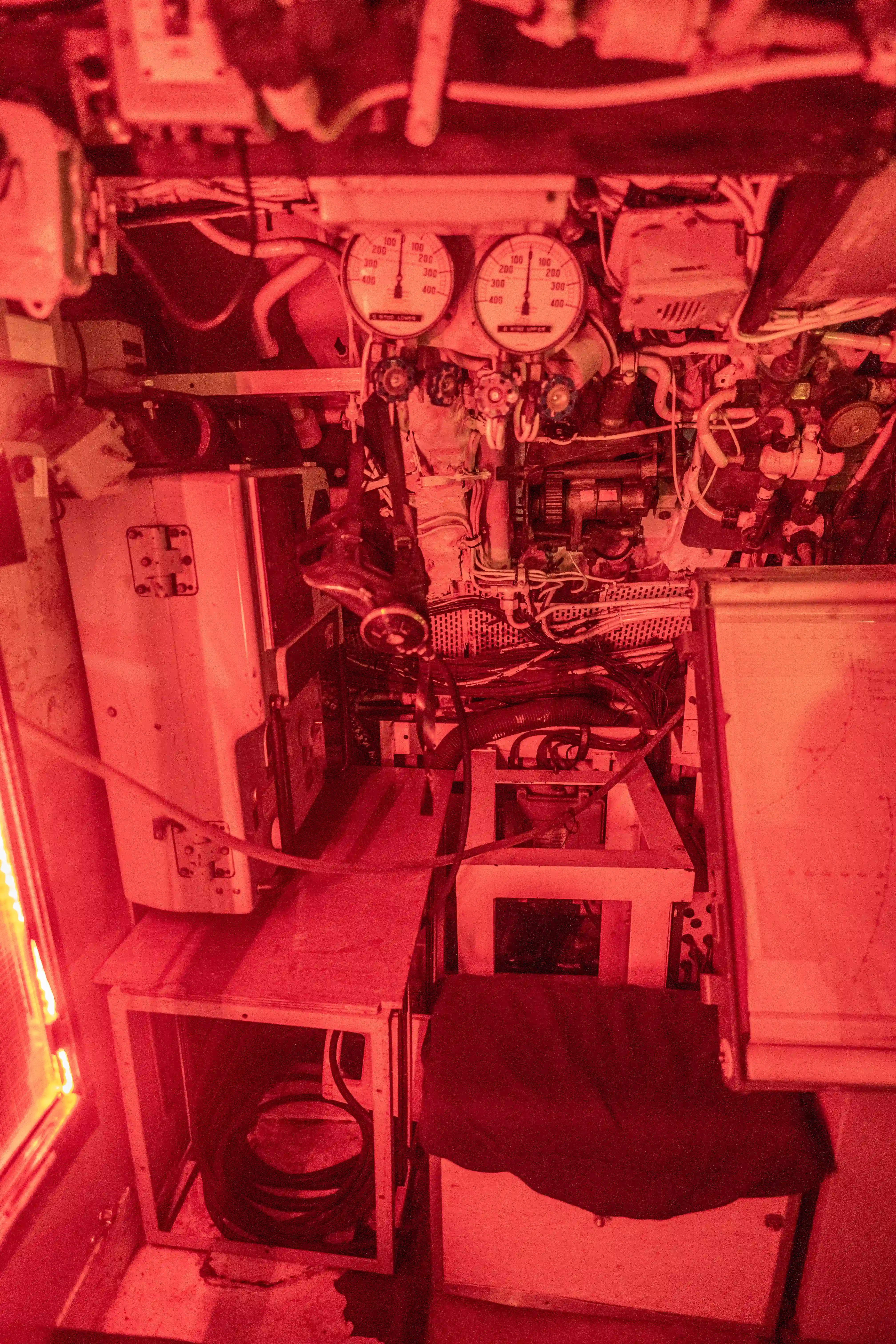 The image depicts a complex control room or engine room with various mechanical components, gauges, and wiring. The lighting is red, creating a distinct atmosphere. There are two prominent gauges at the top center, and numerous pipes and cables are visible throughout the space. The room appears to be part of a larger mechanical or industrial system, possibly within a submarine or ship.
