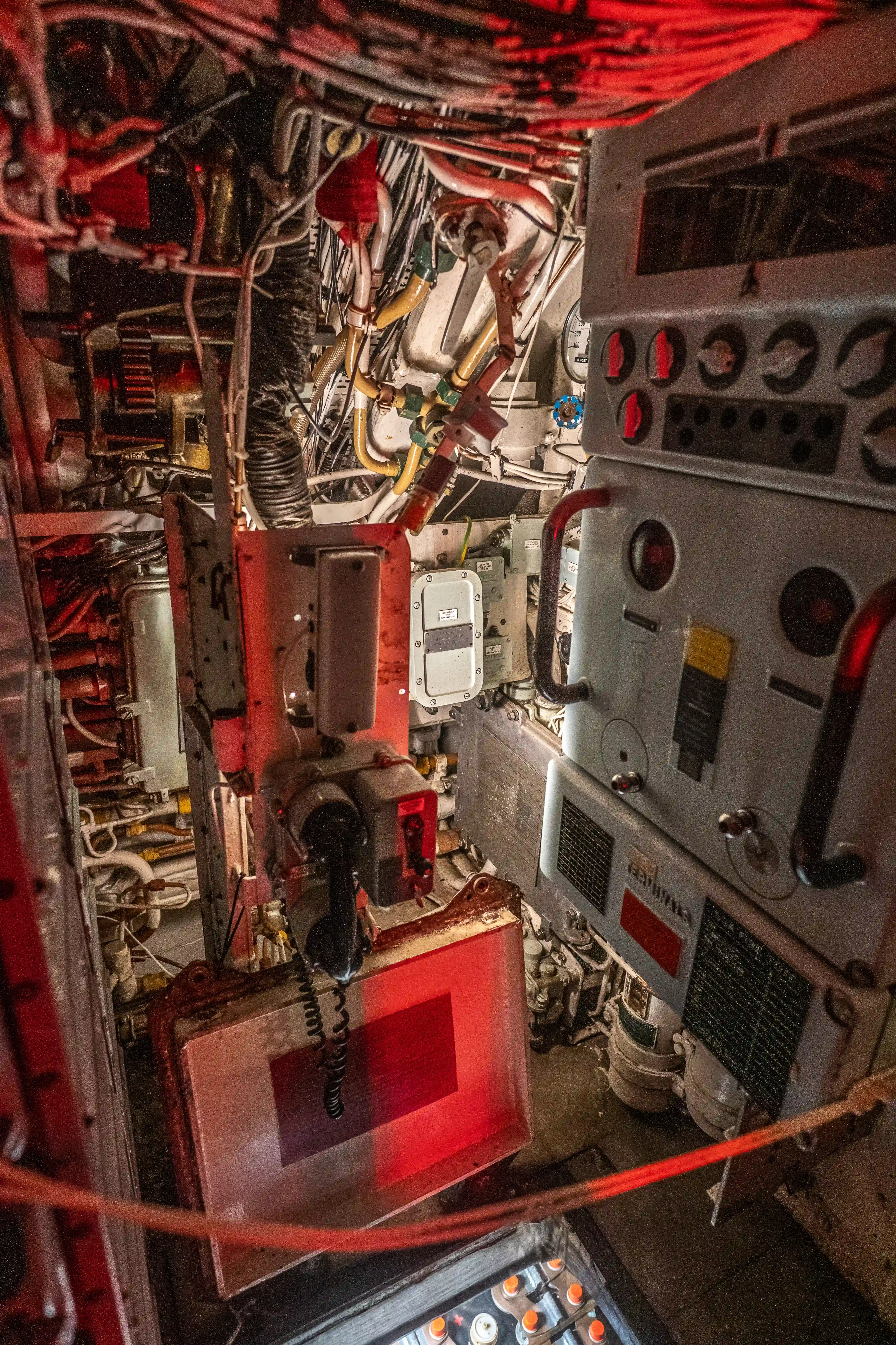 The image depicts the interior of a complex mechanical or industrial space, likely the control room of a submarine or a similar vessel. It features an array of intricate machinery, control panels, and numerous wires and cables. The lighting is predominantly red, which is typical for environments requiring low-light visibility. Various gauges, switches, and equipment are visible, indicating a highly functional and technical area.