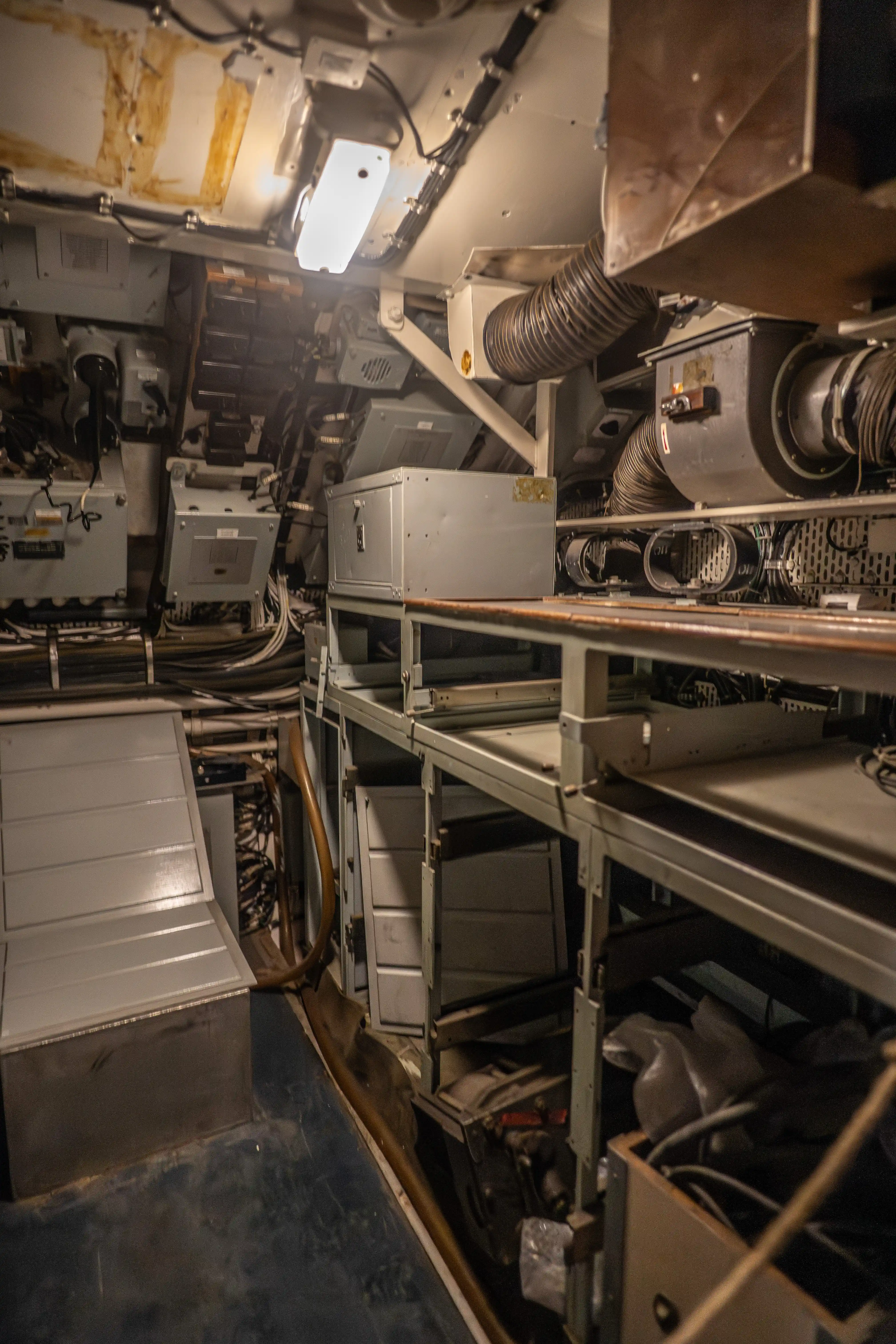 The image depicts the interior of a confined, industrial space, likely a mechanical or electrical room. It is filled with various equipment, including metal cabinets, ventilation ducts, and numerous cables and wires. The area is well-lit with overhead fluorescent lighting. The environment appears to be utilitarian and functional, designed for operational efficiency rather than comfort.