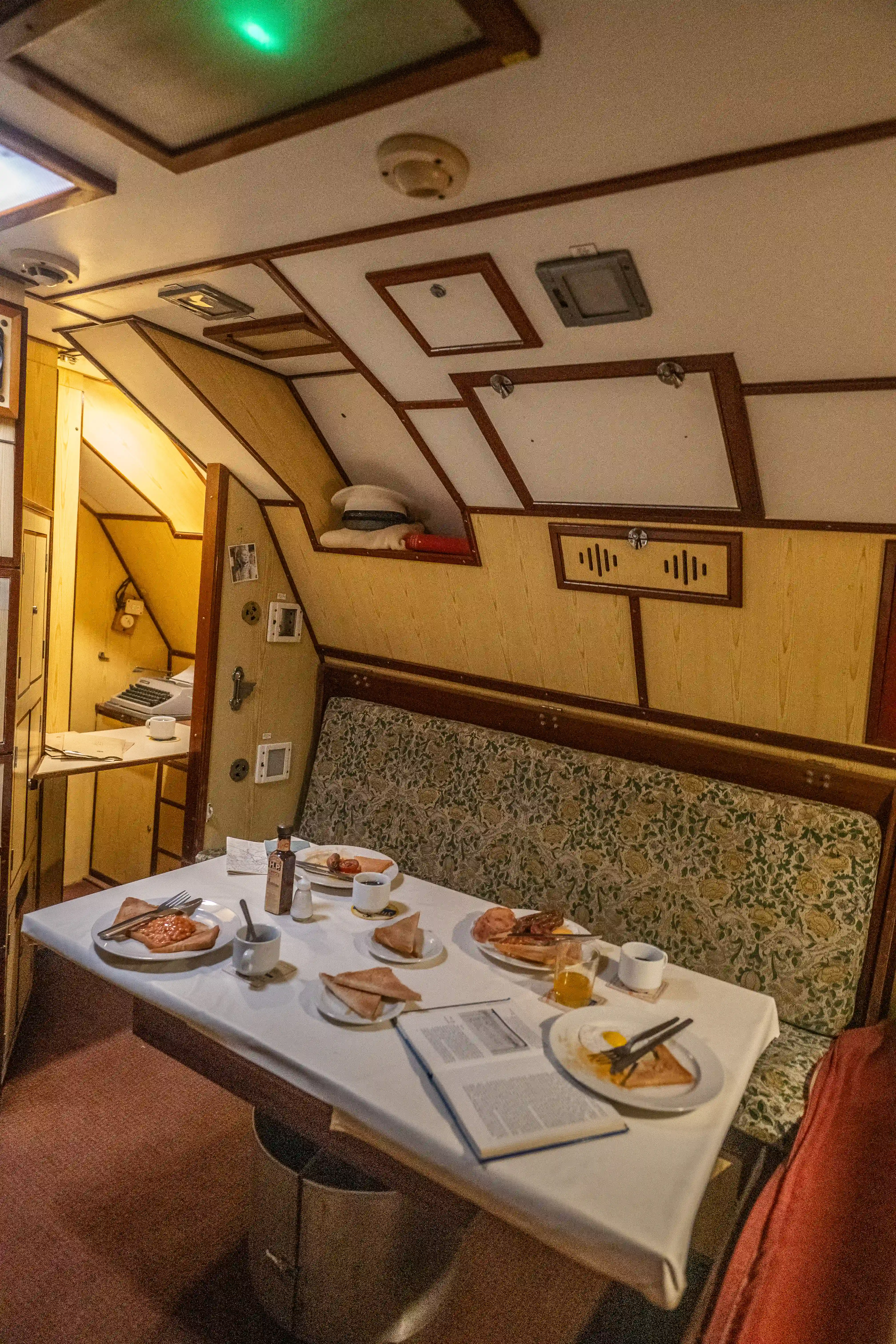 The image depicts a vintage dining area, likely within an aircraft or train. The table is set with various breakfast items including plates of food, cutlery, and beverages. The decor features wooden paneling, a patterned bench seat, and overhead compartments. There is a small book or menu on the table, and a hat is visible on an upper shelf. The setting suggests a retro or historical ambiance.