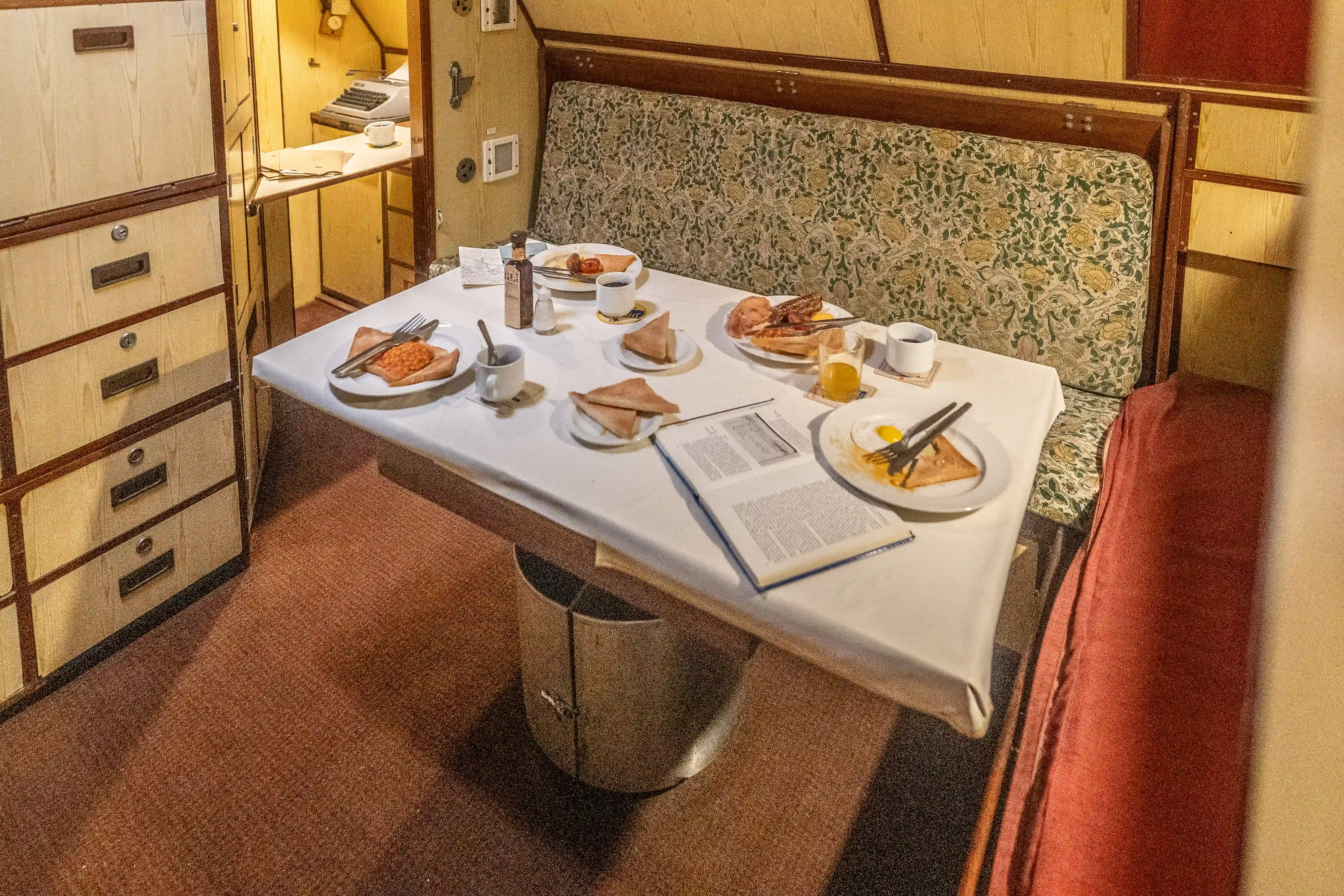The image depicts a small dining area, likely within a train compartment. The table is set with various breakfast items including toast, eggs, and beverages. There are also condiments, cutlery, and an open book or menu on the table. The seating area features a patterned bench, and there are storage drawers and a typewriter visible in the background.