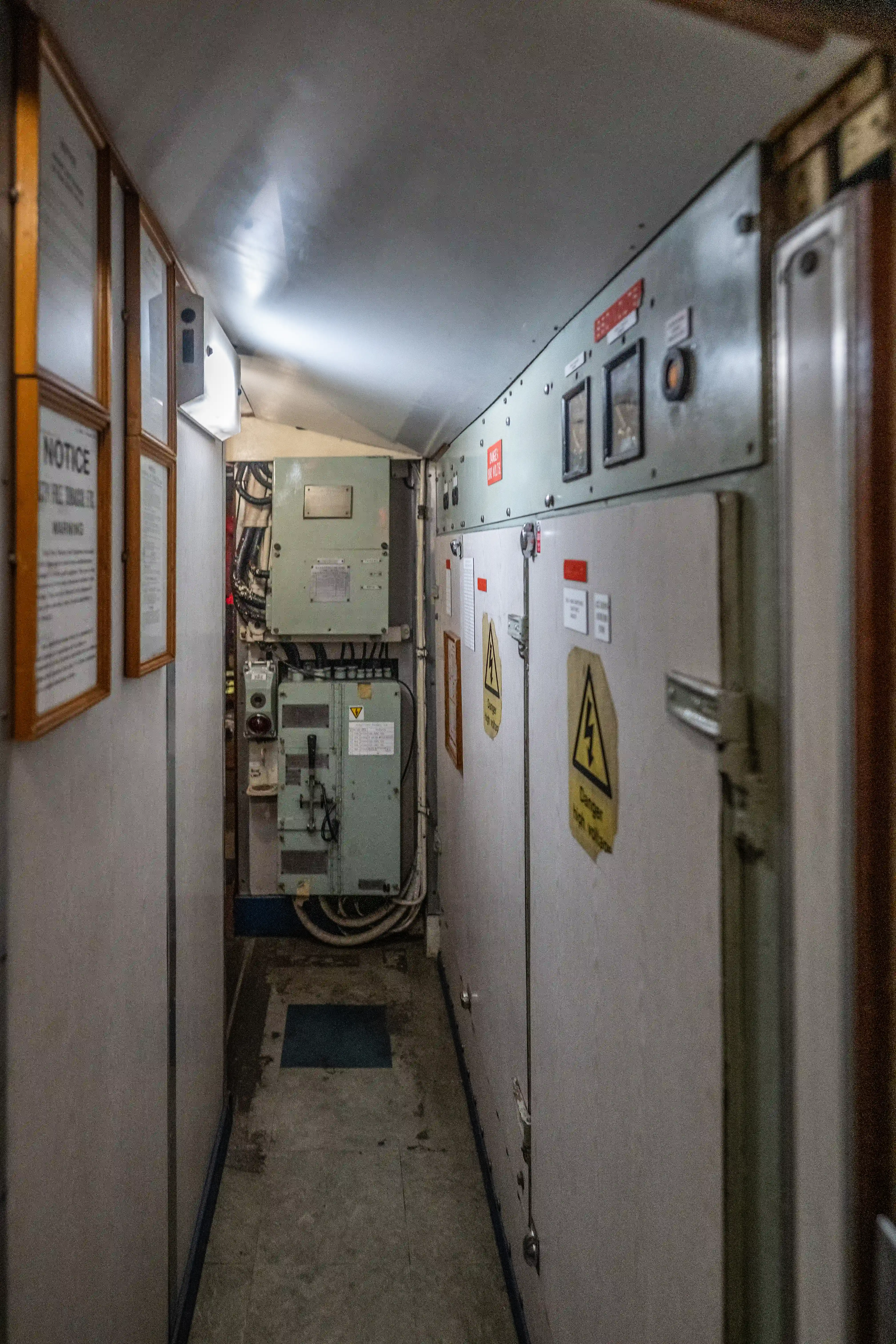 The image depicts a narrow corridor with various electrical panels and equipment. There are warning signs indicating high voltage and caution. The walls are lined with framed notices and documents. The area appears to be a technical or maintenance section, possibly on a ship or industrial facility.