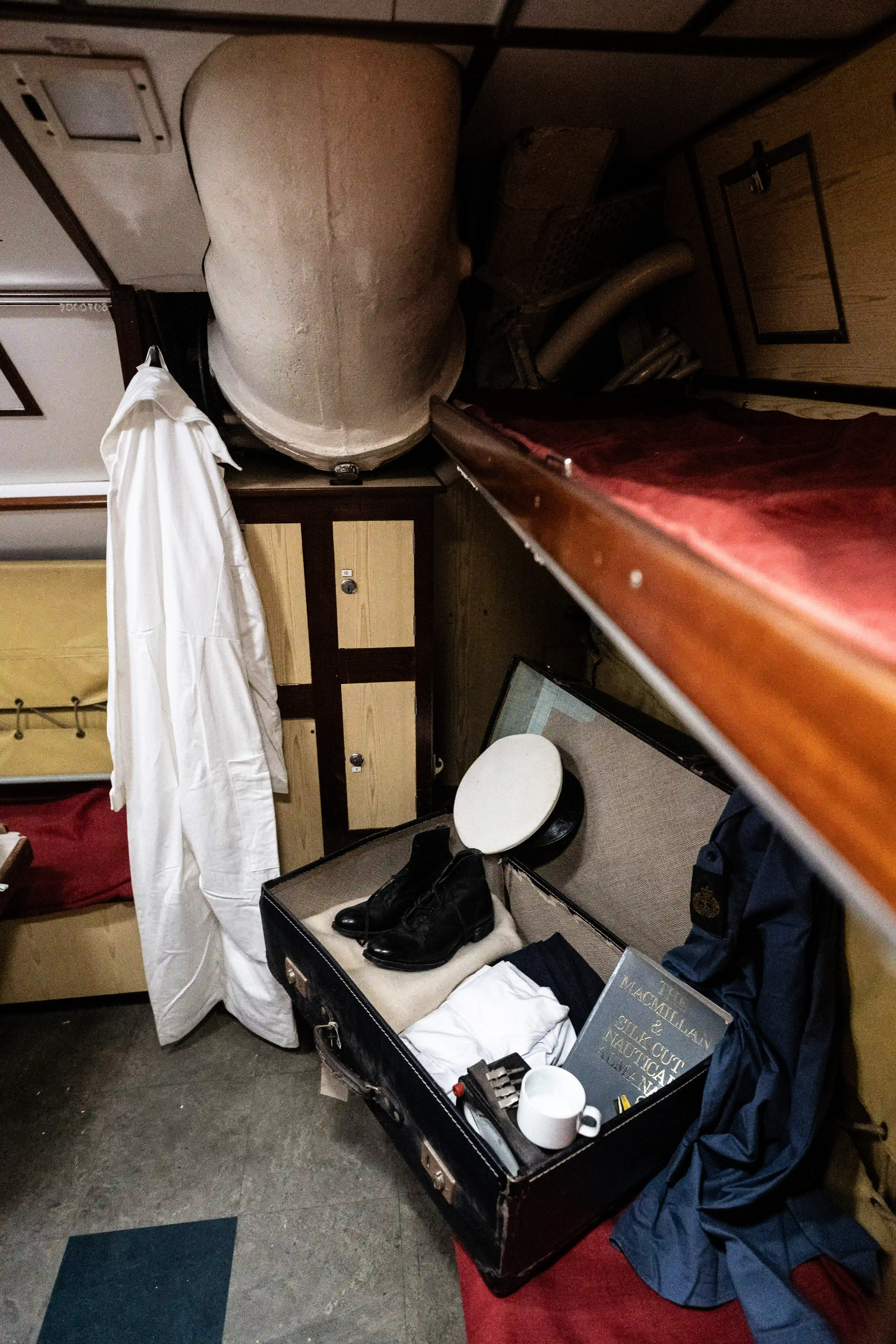 The image depicts a small, confined space, likely a cabin or a ship's quarters. An open suitcase on a lower bunk contains various items, including a pair of black shoes, a white hat, a book titled 'The MacMillan & Silk Cut Nautical Almanac,' and a cup. A white garment hangs on a hook near the suitcase. The setting suggests a historical or vintage theme, possibly from a maritime context.