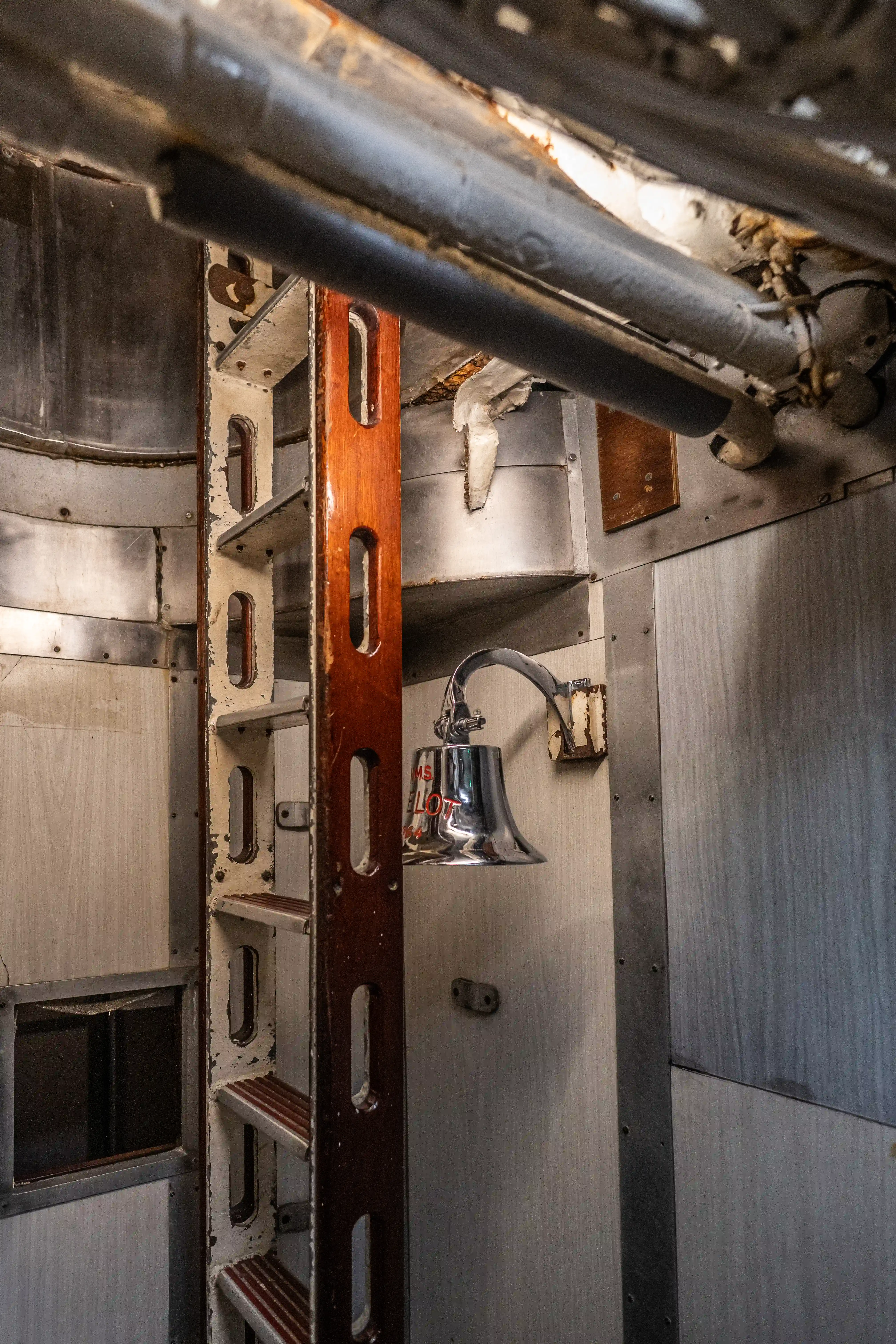 The image depicts an interior view of what appears to be an industrial or marine setting. A vertical ladder is prominently featured, leading upwards through a series of openings in the structure. A metallic bell is mounted on the wall to the right of the ladder, with some text visible on it. The surroundings are primarily made of metal, with visible pipes and structural elements, indicating a utilitarian environment, possibly within a ship or submarine.