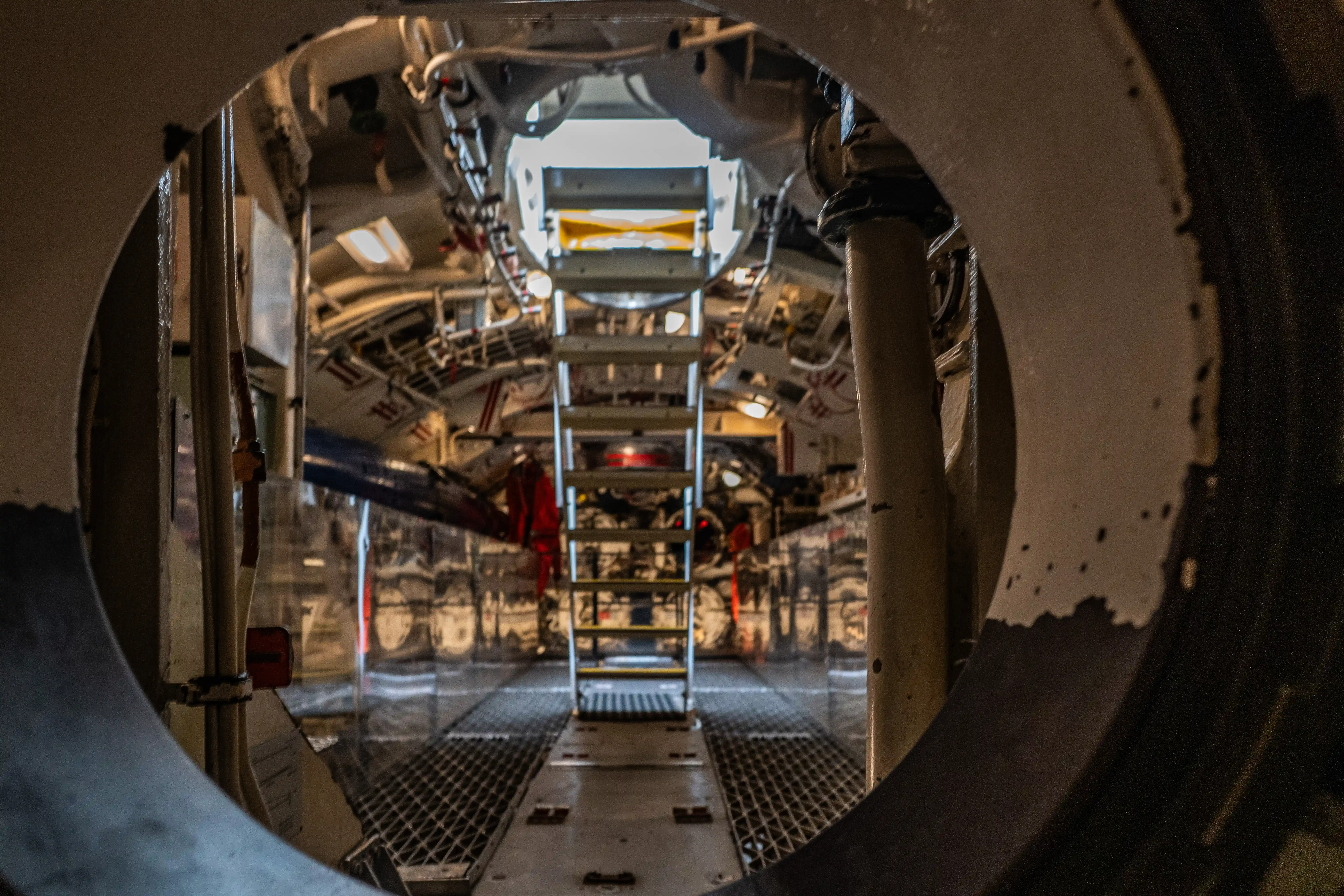 The image depicts the interior of a submarine viewed through a circular hatch. The scene includes various mechanical components, pipes, and equipment typical of a submarine's engine room or similar compartment. A ladder is visible leading to different levels, and there are various tools and safety gear, including red protective suits. The lighting is industrial, and the overall environment appears to be tightly packed and highly functional.