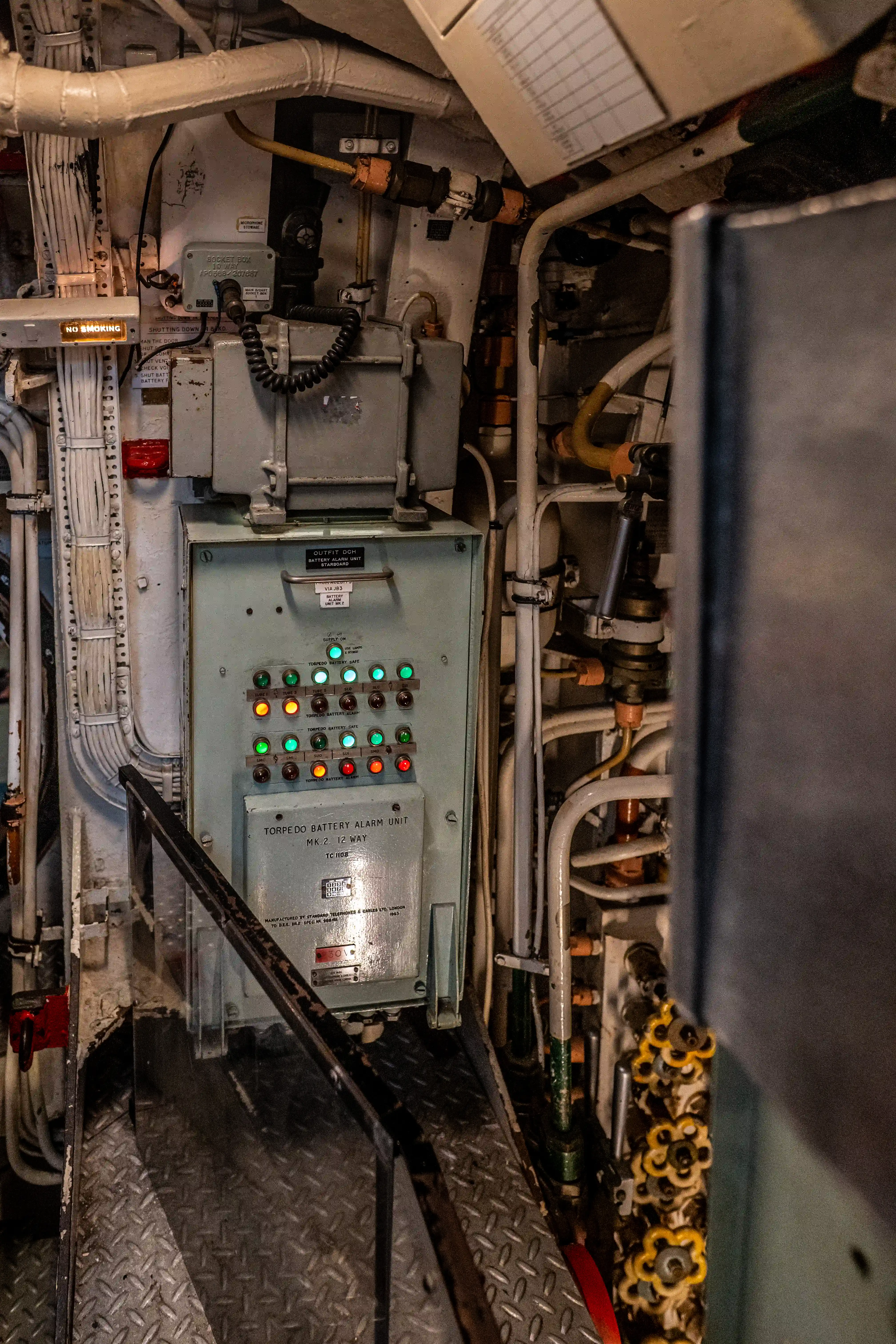 The image depicts a complex control panel and machinery setup, likely within an industrial or naval environment. The central focus is on a control unit labeled 'Torpedo Battery Alarm Unit,' featuring multiple buttons and indicators. Surrounding the control unit are various pipes, cables, and structural elements, suggesting a densely packed and highly functional space. The area appears to be well-maintained, with clear labeling and organized components. The overall setting indicates a technical and operational area, possibly within a submarine or a similar vessel.