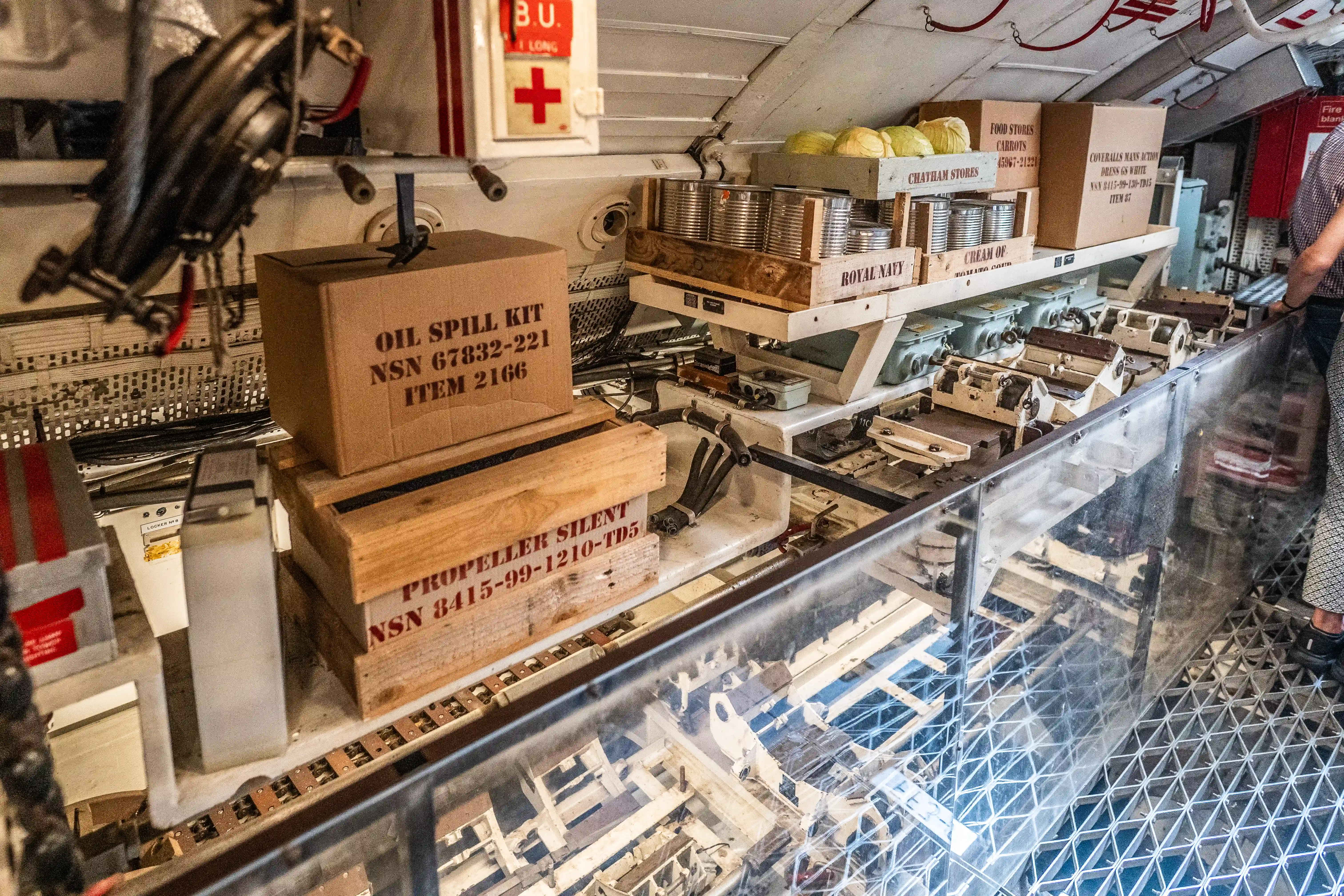 The image depicts a storage area, likely on a naval vessel or submarine, containing various supplies and equipment. Notable items include an 'Oil Spill Kit' and a 'Propeller Silent' component, both with specific NSN (National Stock Number) and item numbers. There are also boxes labeled for food stores, including 'Cream of Royal Navy,' and other equipment secured on shelves. The area is organized with storage bins and racks, and a person is visible on the right side of the image, suggesting maintenance or inspection activities.