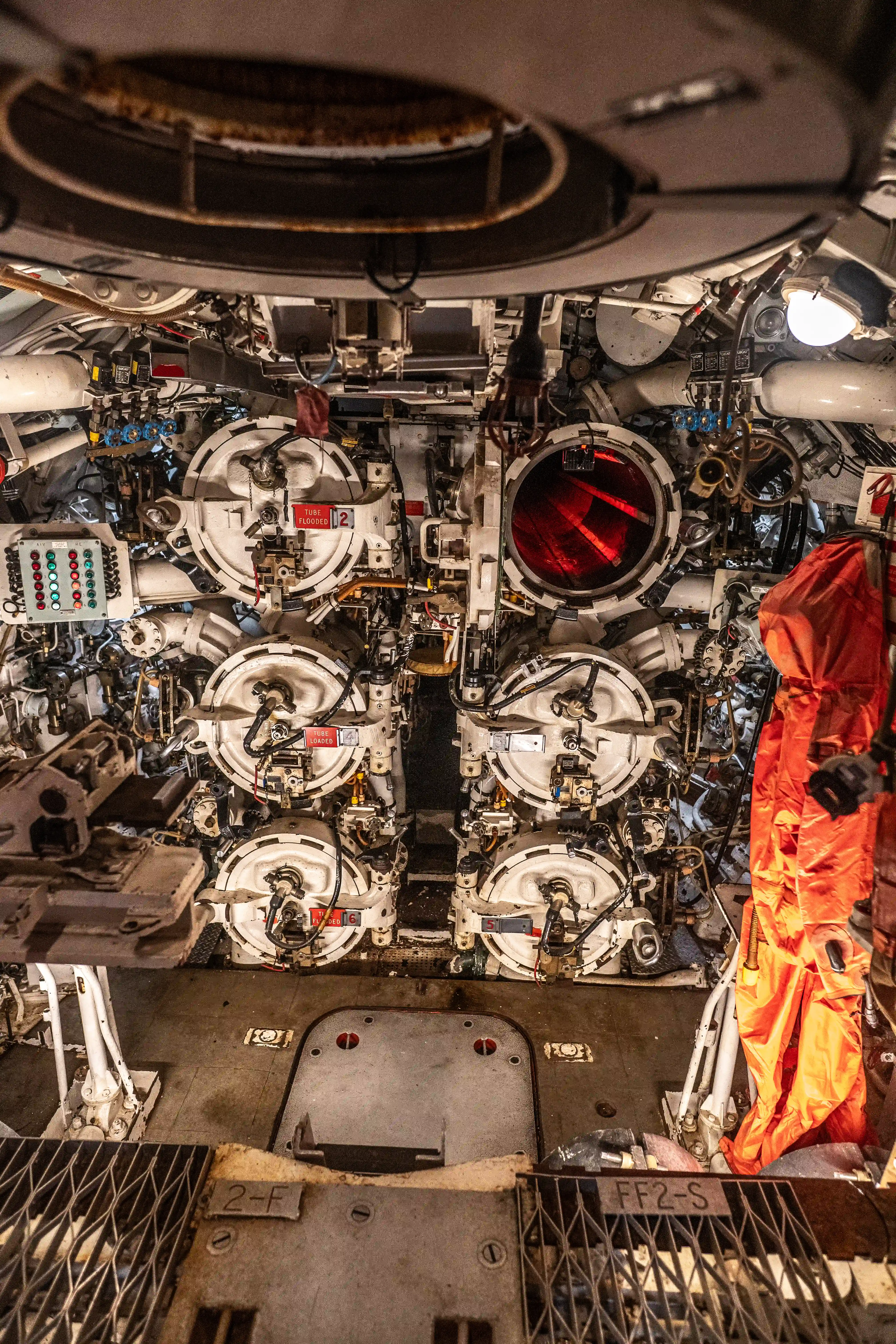 he image depicts the interior of a submarine, specifically the torpedo room. It shows multiple torpedo tubes, control panels, and various mechanical components. The room is filled with complex equipment and machinery essential for the operation and launching of torpedoes. The environment appears to be tightly packed with intricate details and technical instruments.