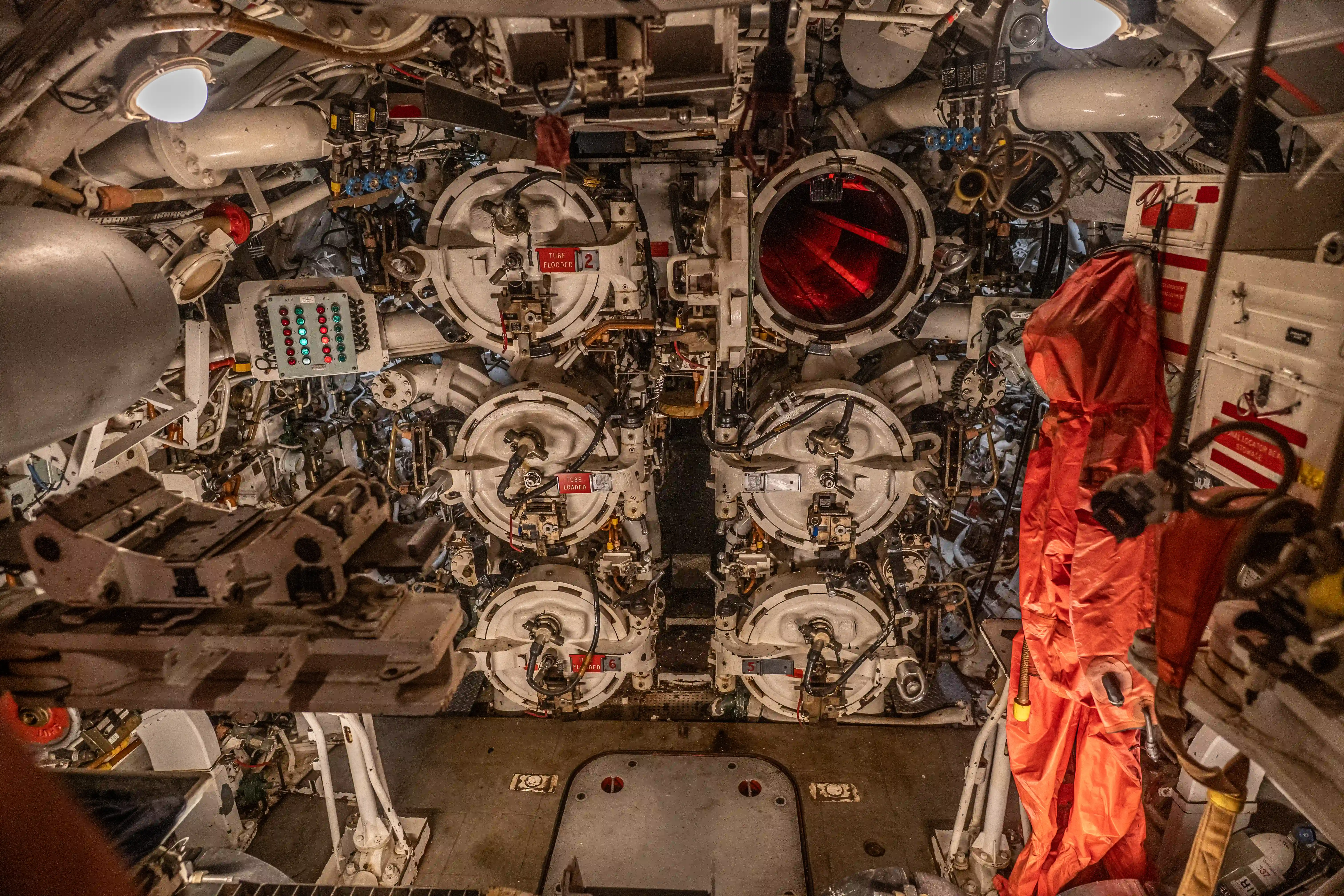 The image depicts the interior of a submarine, specifically the torpedo room. This area is densely packed with mechanical equipment, including several torpedo tubes, control panels, and various machinery. The room is illuminated by overhead lights, and there are red covers and signs indicating safety and operational instructions. The complex arrangement of equipment highlights the intricate and highly technical environment necessary for submarine operations.