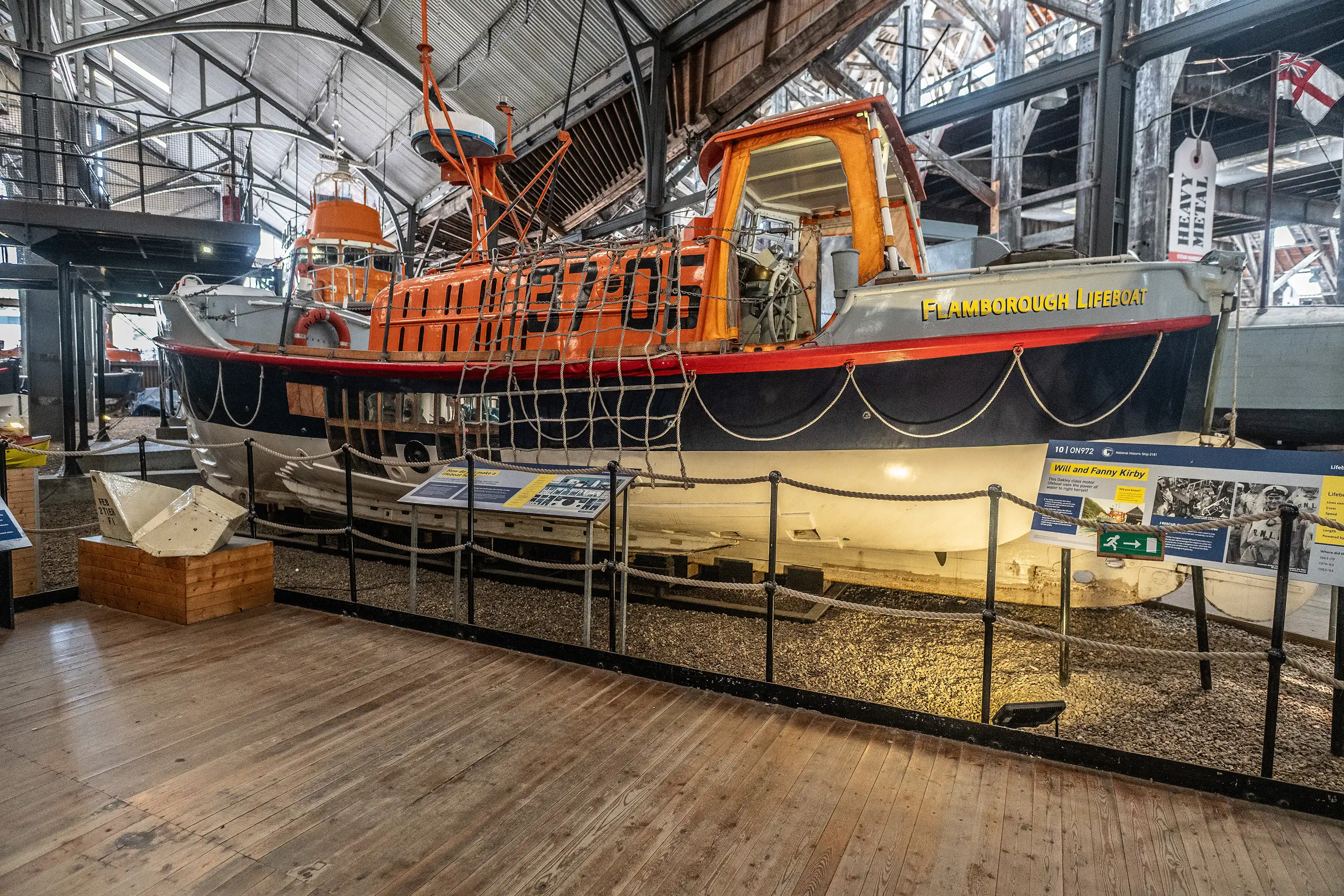 A historic orange and white lifeboat named Flamborough Lifeboat is displayed indoors on a gravel floor behind a rope barrier, with wooden floorboards in the foreground and museum infrastructure in the background.