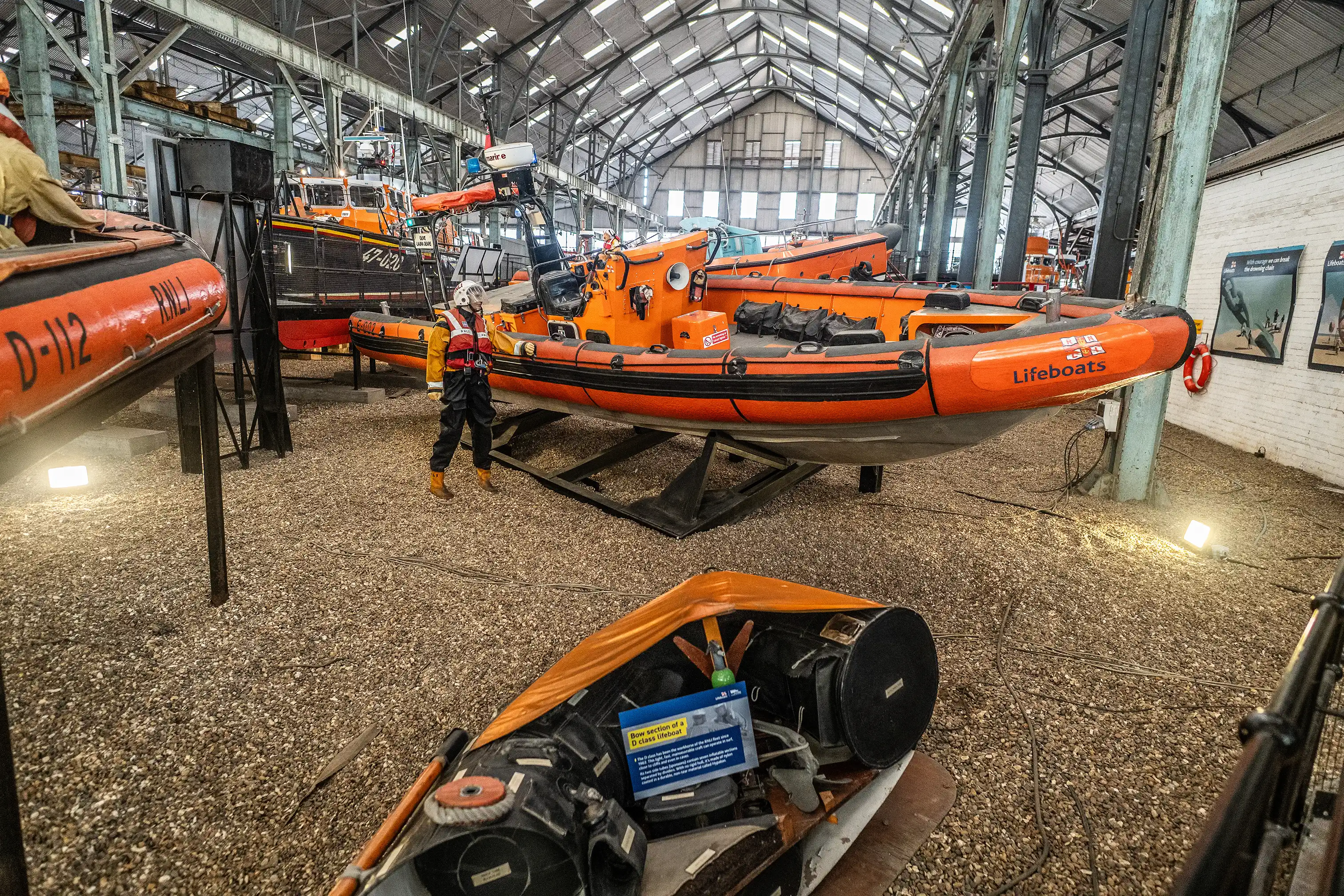 A museum display of orange RNLI lifeboats, with a mannequin in a life vest and several boats visible inside a large industrial building with a gravel floor.
