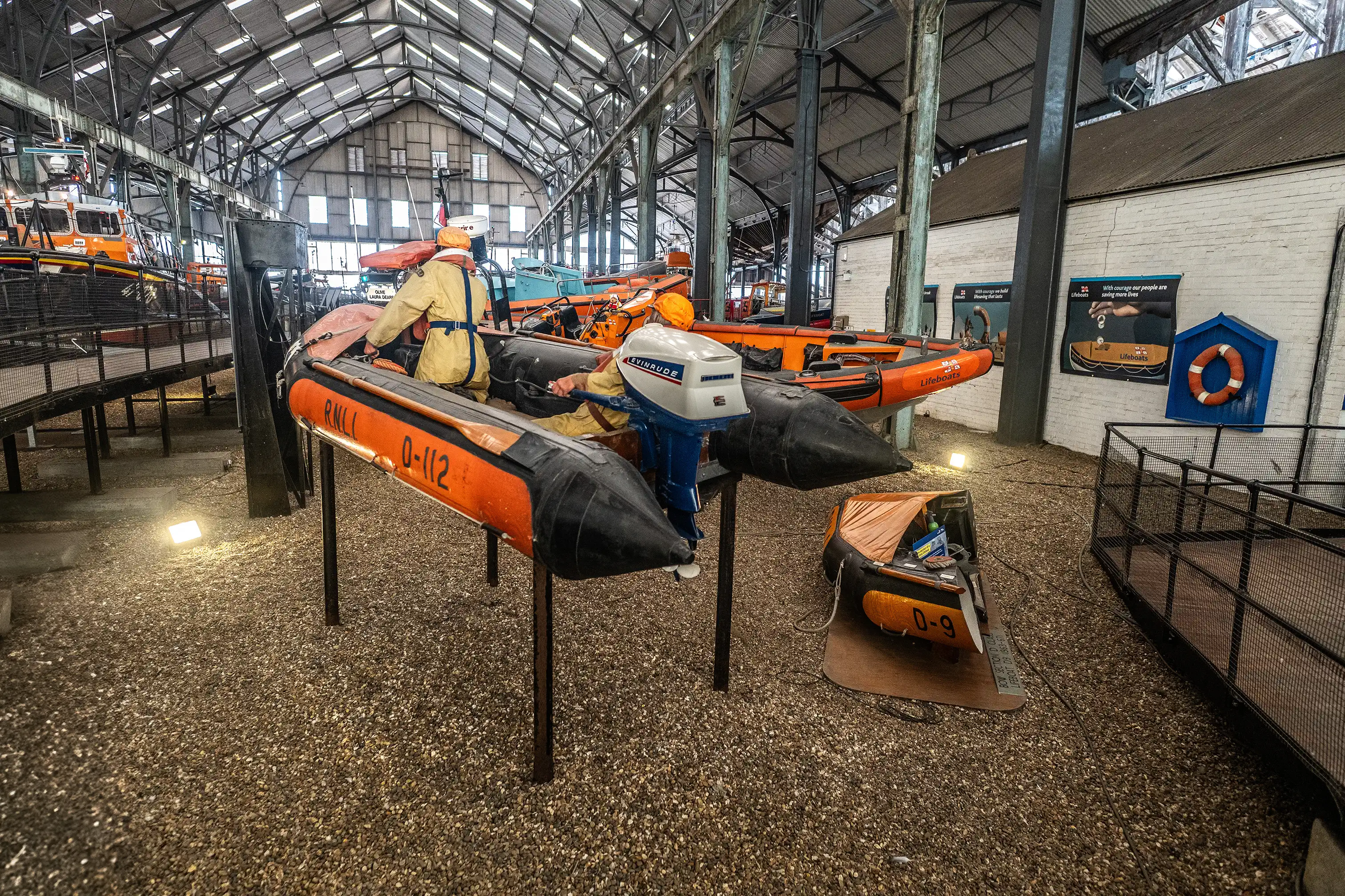 Two RNLI inflatable lifeboats are displayed indoors on a gravel floor. One boat, labelled RNLI 0-112, has mannequins in survival suits inside and is elevated on metal stands. A smaller orange boat is on the floor to the right. The museum setting features a high, industrial roof and white brick walls with framed posters and a lifebuoy.