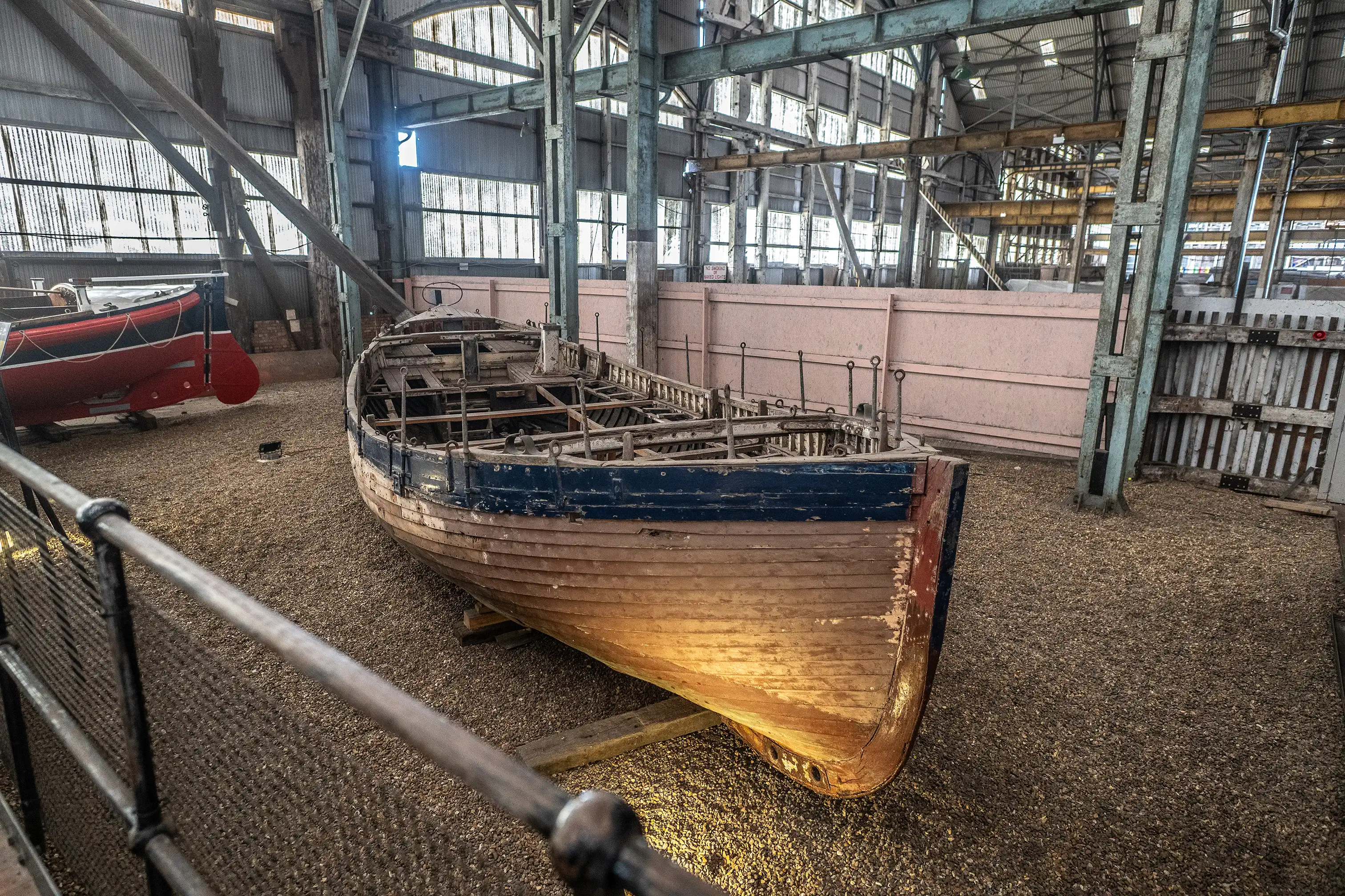 A close-up of the front of a wooden boat with a blue stripe along its upper edge, sitting inside a dimly lit workshop. The boat is made of weathered wooden planks and has exposed internal framing.