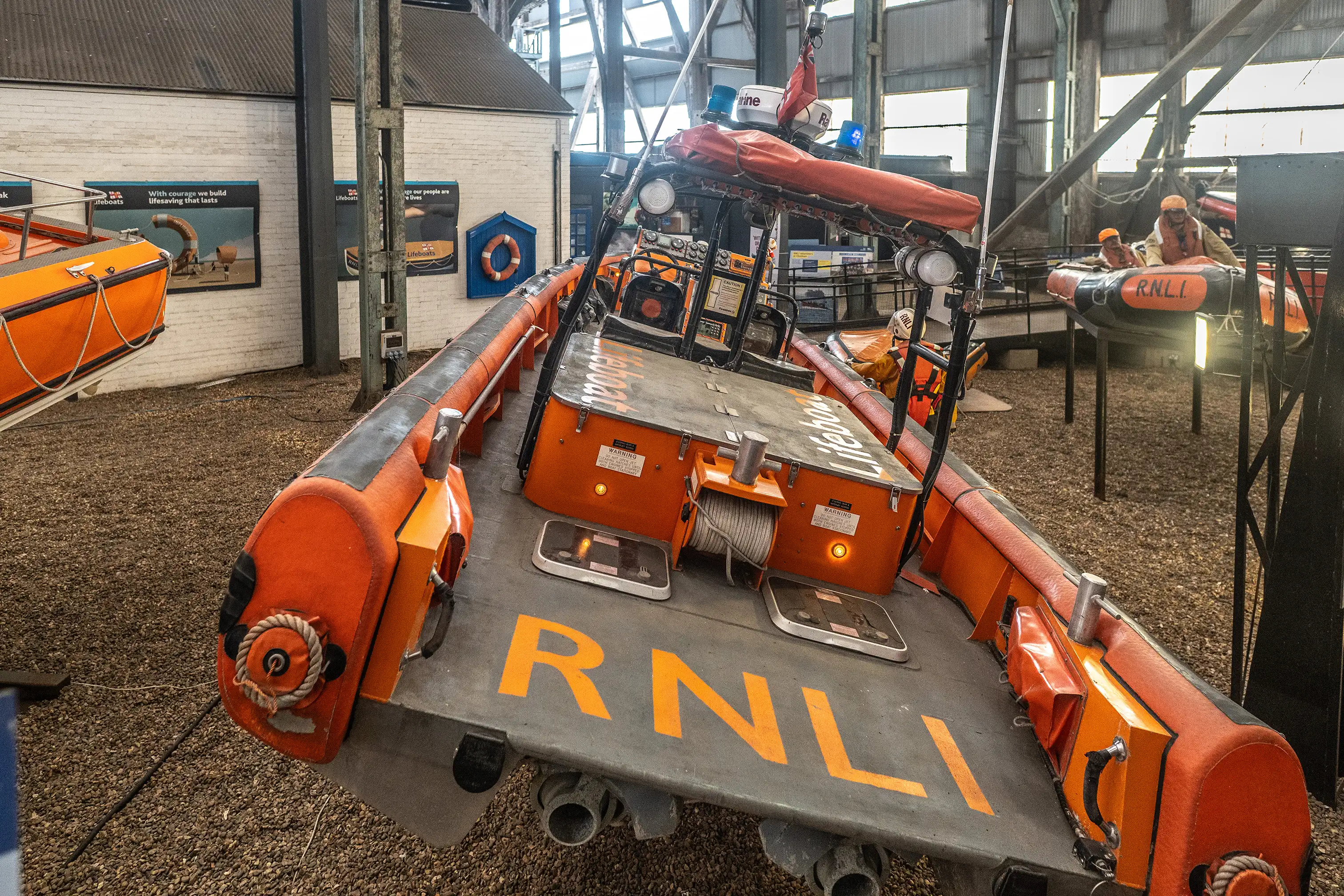 Close-up of the rear deck of an orange and grey RNLI lifeboat, showing the RNLI logo painted in yellow, a rope winch, and metal hatches.