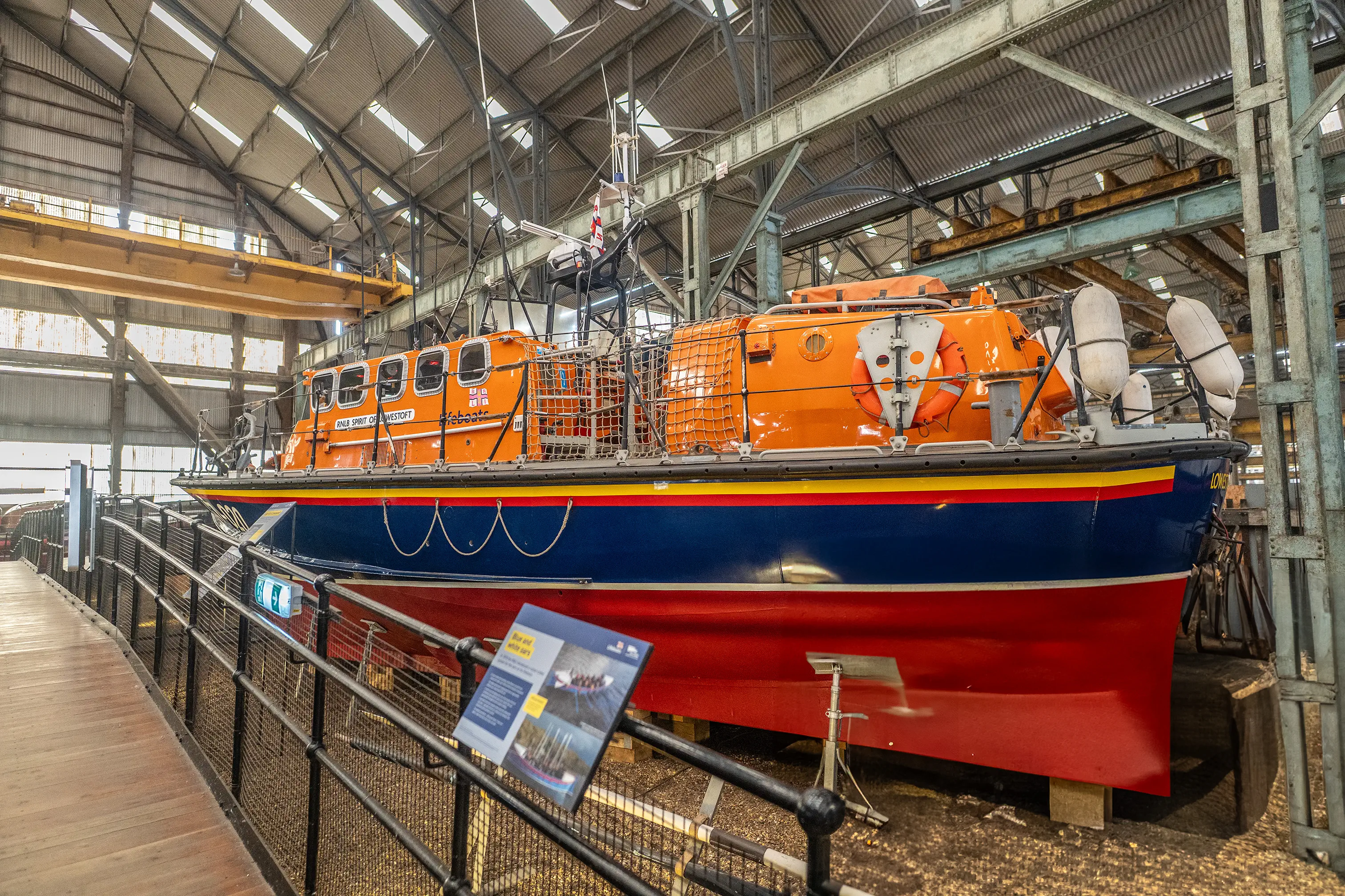  A bright orange RNLI lifeboat with blue and red hull sits in a large industrial shed, viewed from a wooden walkway.