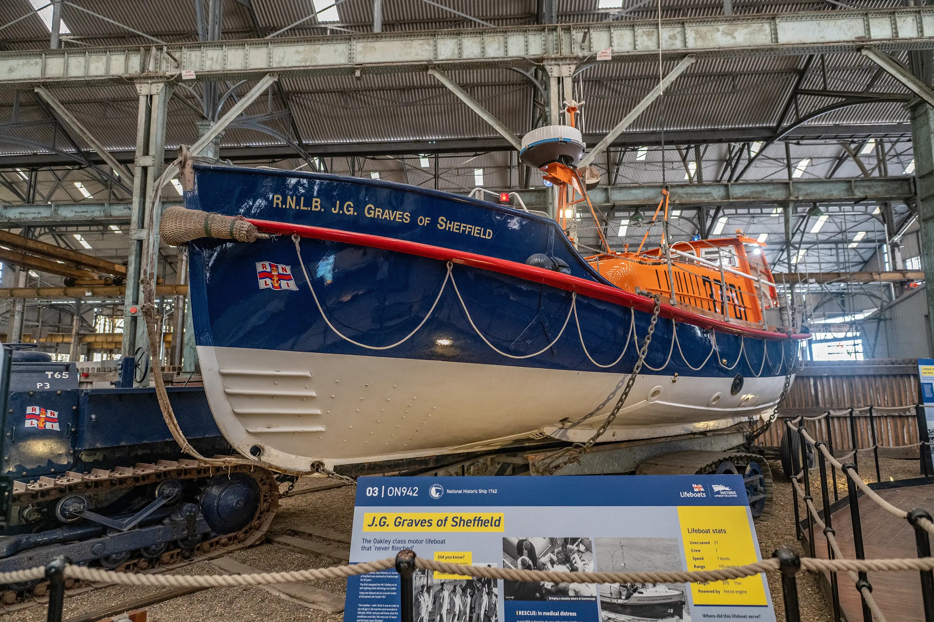 A historic Royal National Lifeboat Institution (RNLI) motor lifeboat, named J.G. Graves of Sheffield, is displayed indoors, possibly in a museum. The lifeboat is blue and white with red trim and an orange canopy. It is mounted on a tracked carriage, suggesting it was launched from land. A sign in the foreground provides information about the lifeboat, including its stats and history. The background shows the industrial-style roof structure of the building.