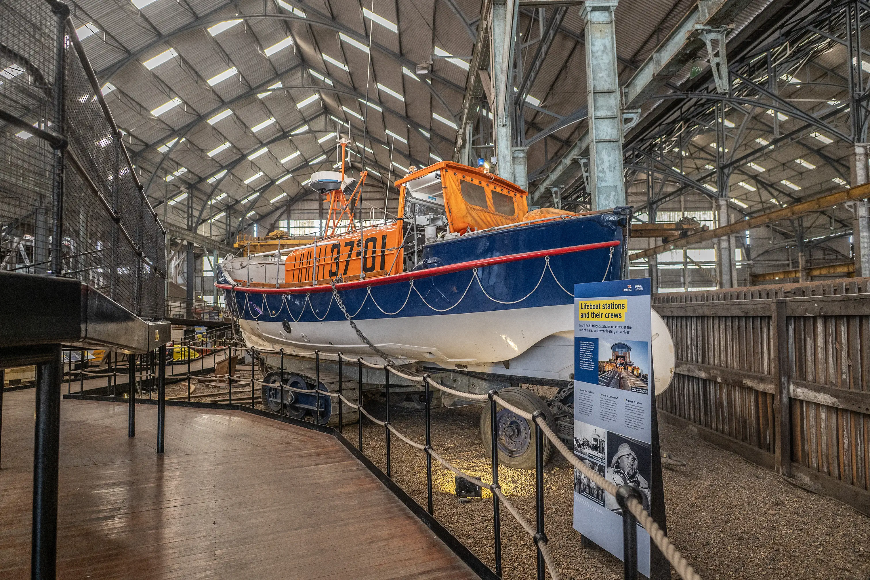 A blue and white RNLI lifeboat with an orange cabin, numbered 37-01, displayed on a wheeled carriage inside a large museum hall with interpretation panels and walkways.