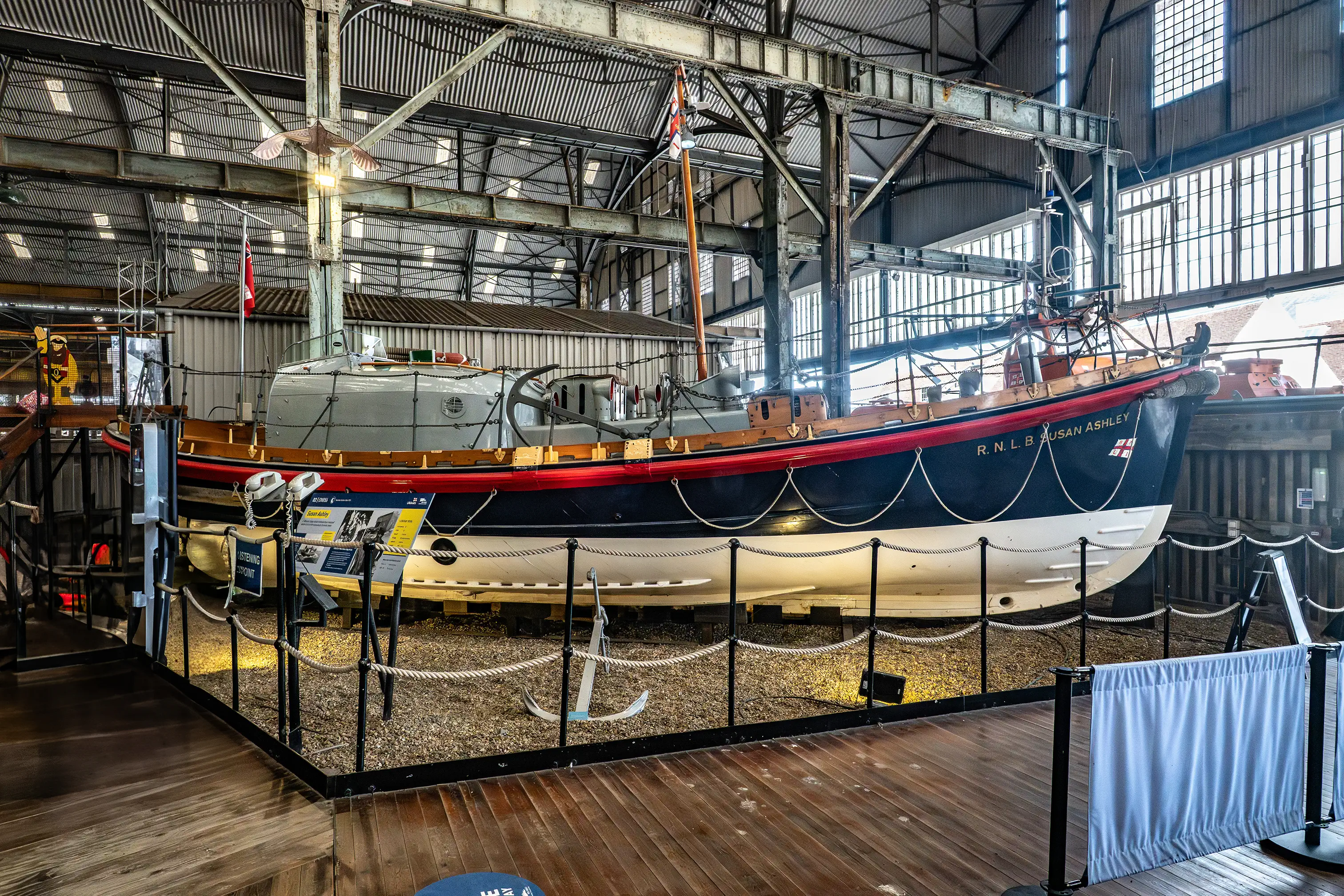 A large blue and white RNLI lifeboat named Susan Ashley displayed on supports inside a maritime museum, with ropes, barriers, and information boards around it.