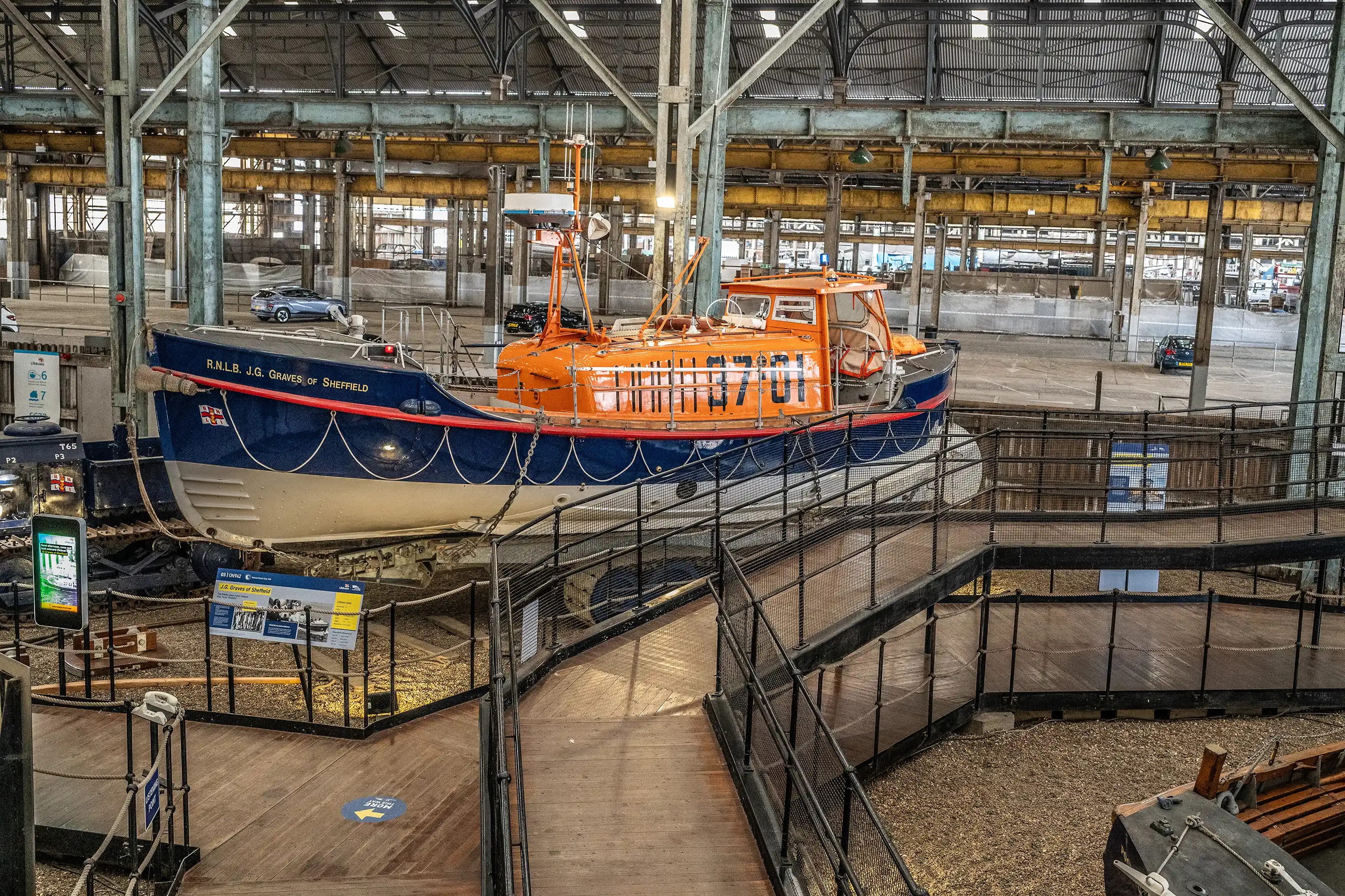 An orange and blue RNLI lifeboat named J.G. Graves of Sheffield is housed inside a large industrial hall, with wooden walkways and a metal railing in the foreground.
