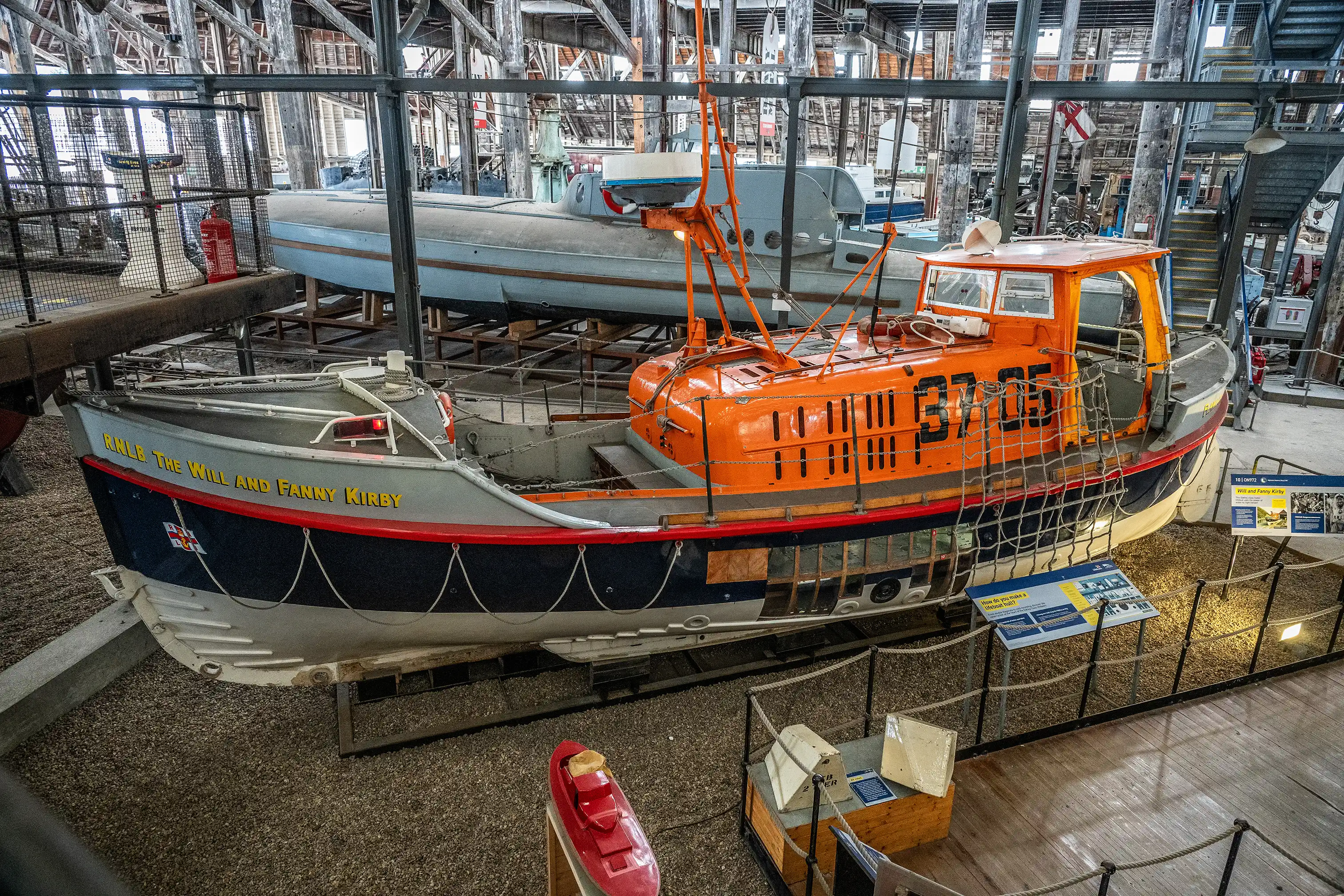 An orange and dark blue RNLI lifeboat, named 'The Will and Fanny Kirby', is displayed indoors in a museum setting, with a larger grey vessel behind it and various informational exhibits around it.