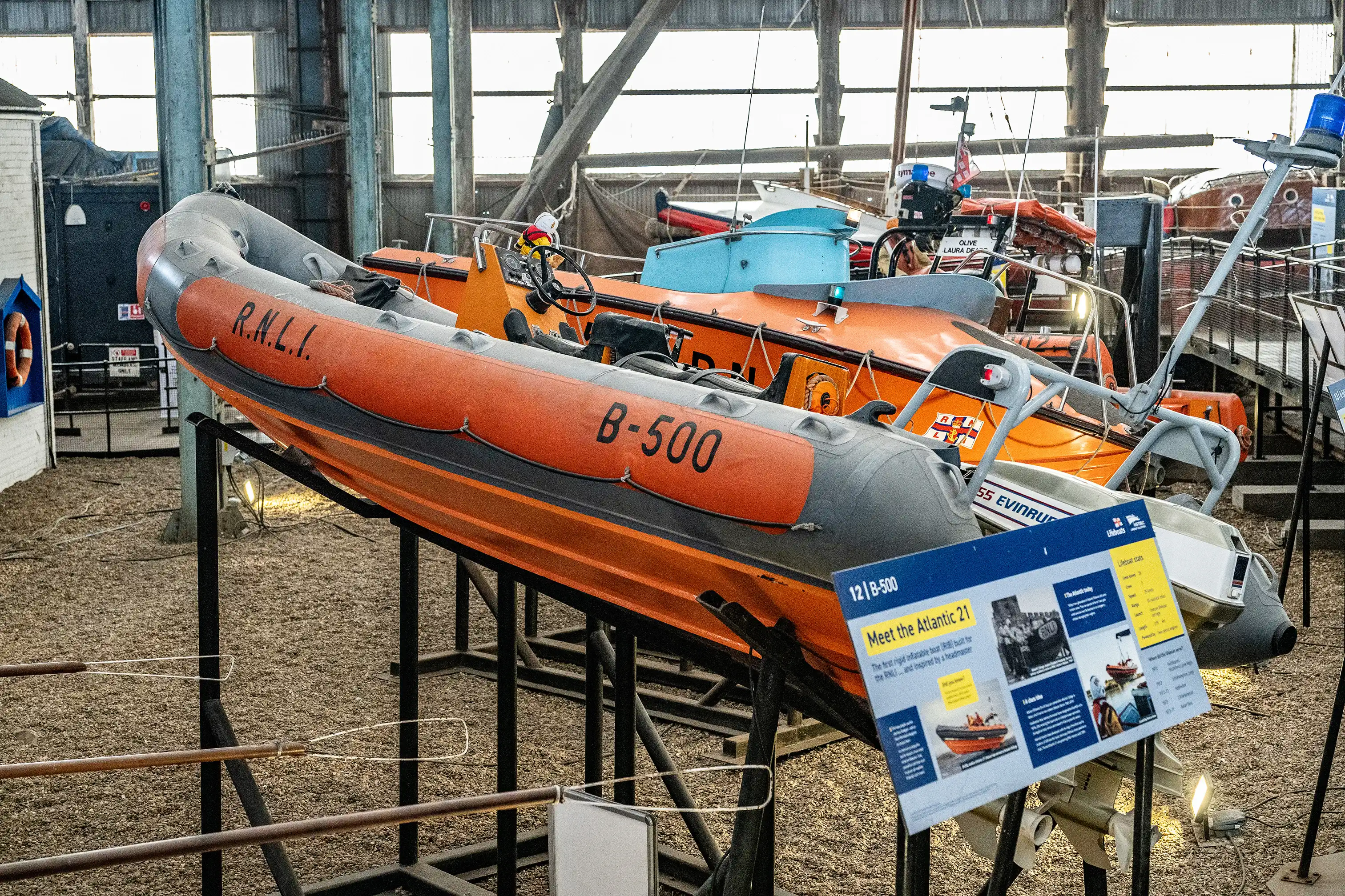 An orange and grey RNLI rigid inflatable lifeboat, marked B-500, is displayed on a stand in a museum-like setting, with other orange lifeboats visible behind it.