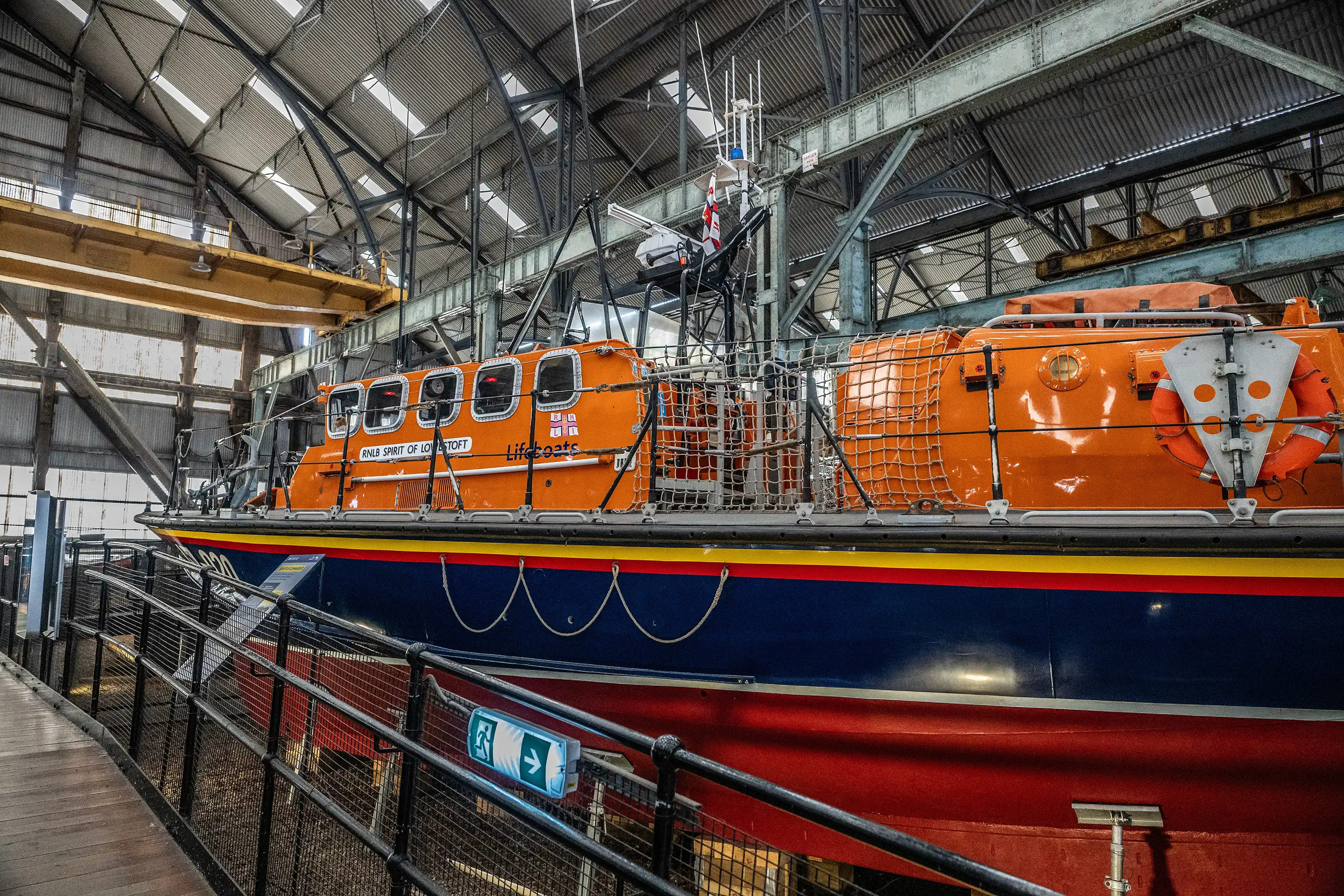 An interior view of a large industrial building housing the orange and blue RNLI (Royal National Lifeboat Institution) lifeboat named Spirit of Lowestoft. The vessel is positioned on a stand, and a wooden walkway with a metal railing and an illuminated green exit sign is visible in the foreground. The building's corrugated metal roof and exposed structural beams are prominent overhead.