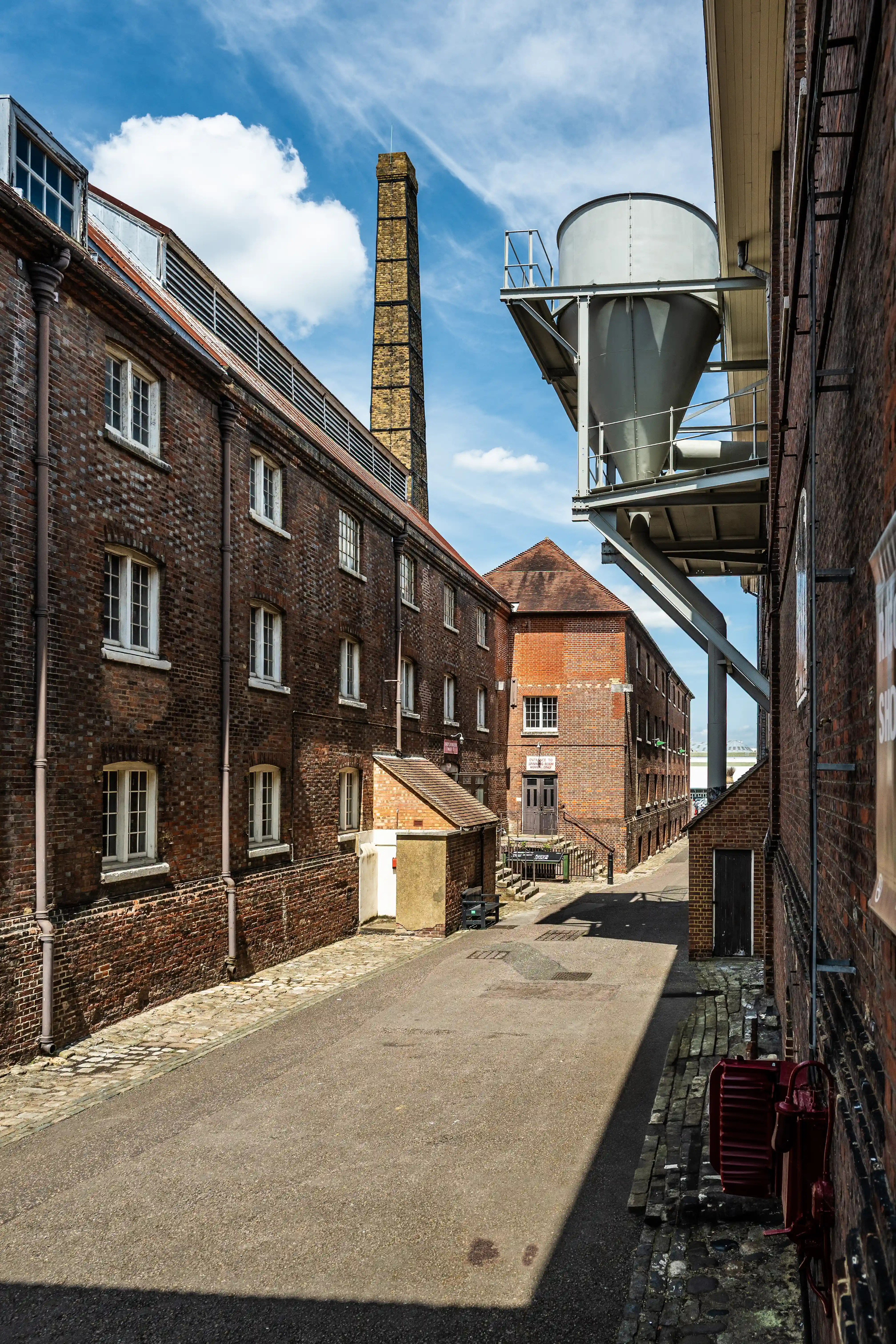 A narrow street lined with red-brick industrial buildings under a blue sky with clouds. A tall chimney and a suspended industrial silo are visible.