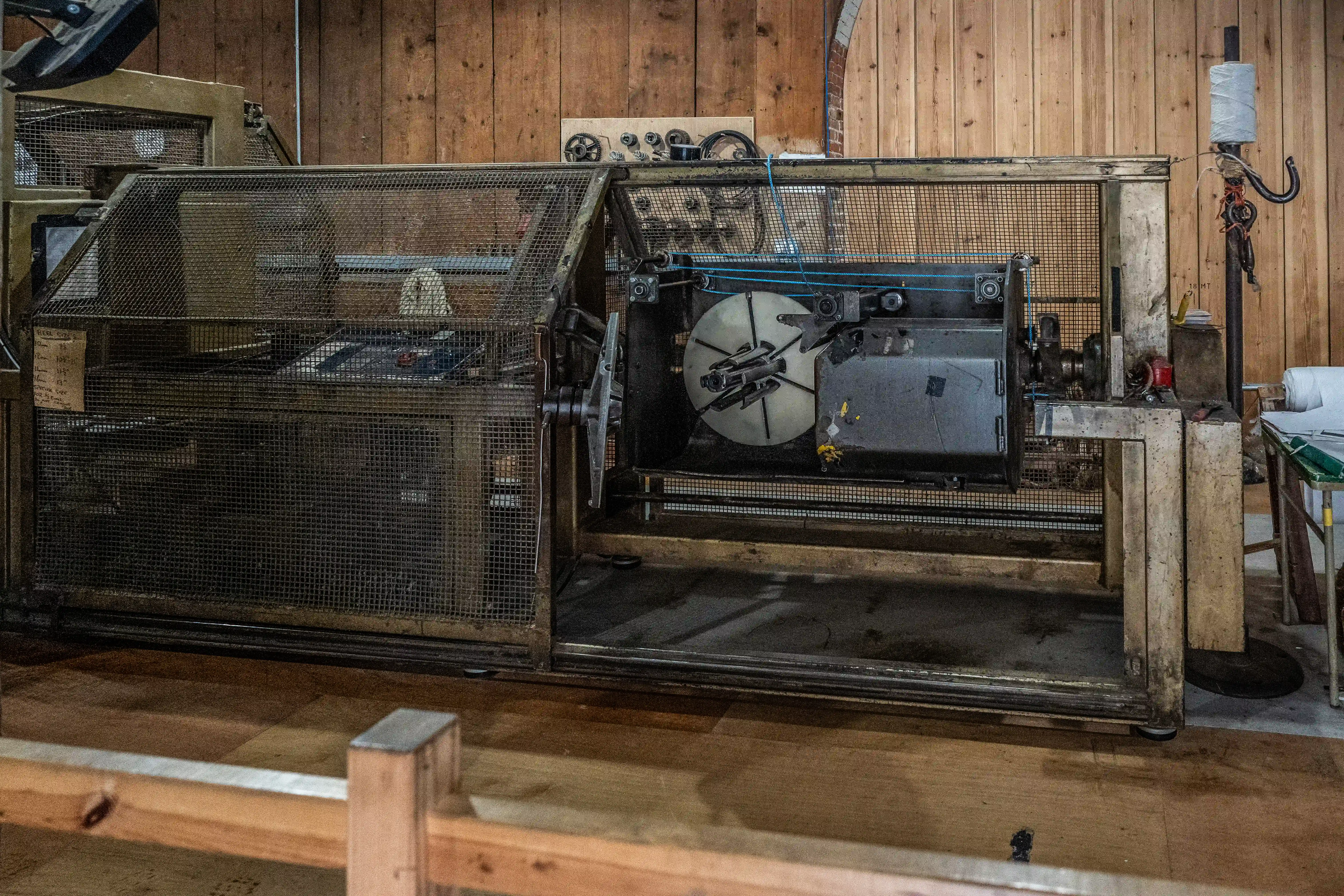 Industrial machine with wire winding mechanism, enclosed in wire mesh, in a workshop setting with wooden walls.