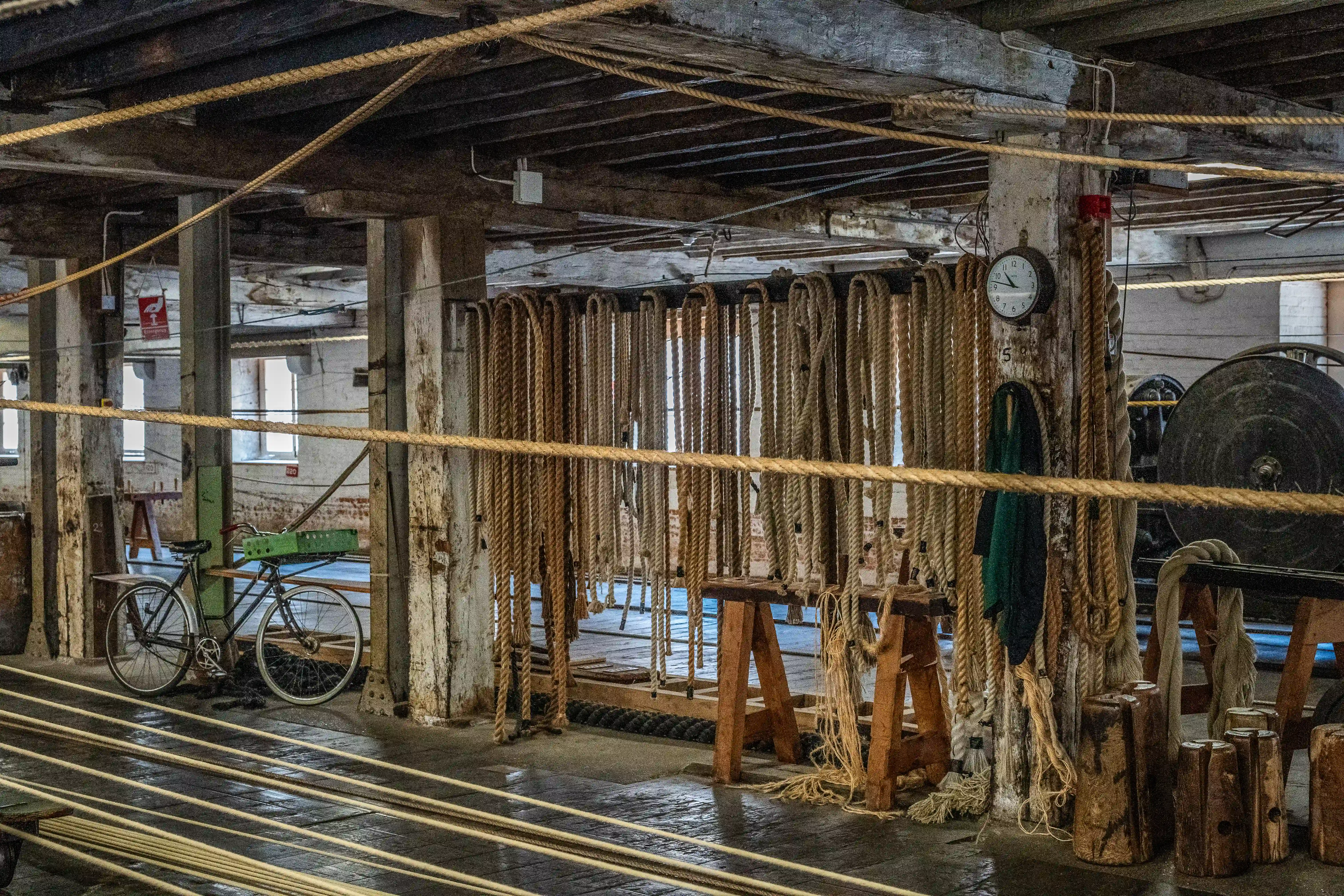 Interior of an old rope-making workshop with many thick, natural fibre ropes hanging and coiled, wooden beams, a vintage bicycle, and a wall clock.