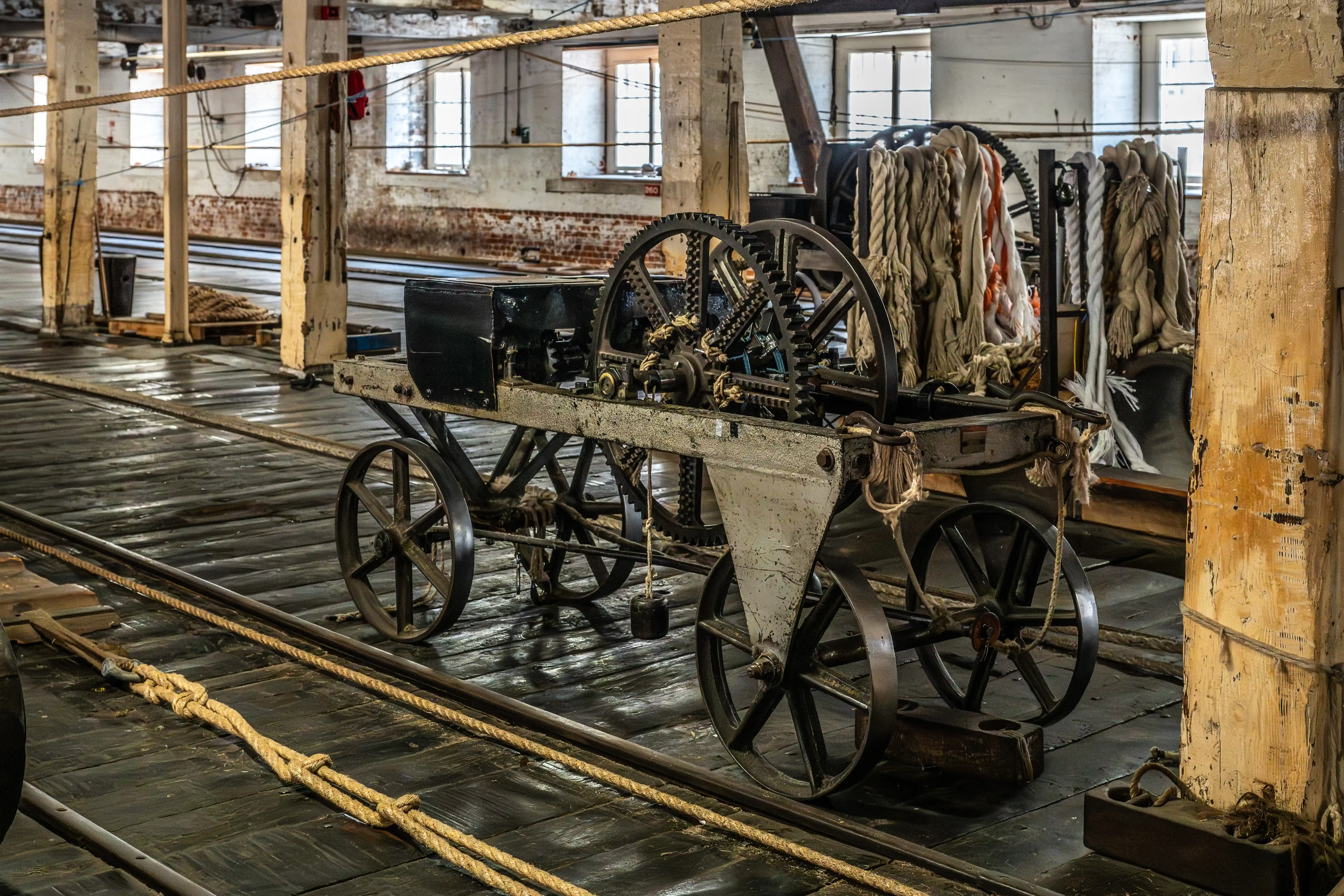 A close-up of a rustic, wheeled industrial machine with large metal gears and spoked wheels, set on a weathered wooden floor amidst coiled ropes and wooden support beams in a dimly lit workshop.