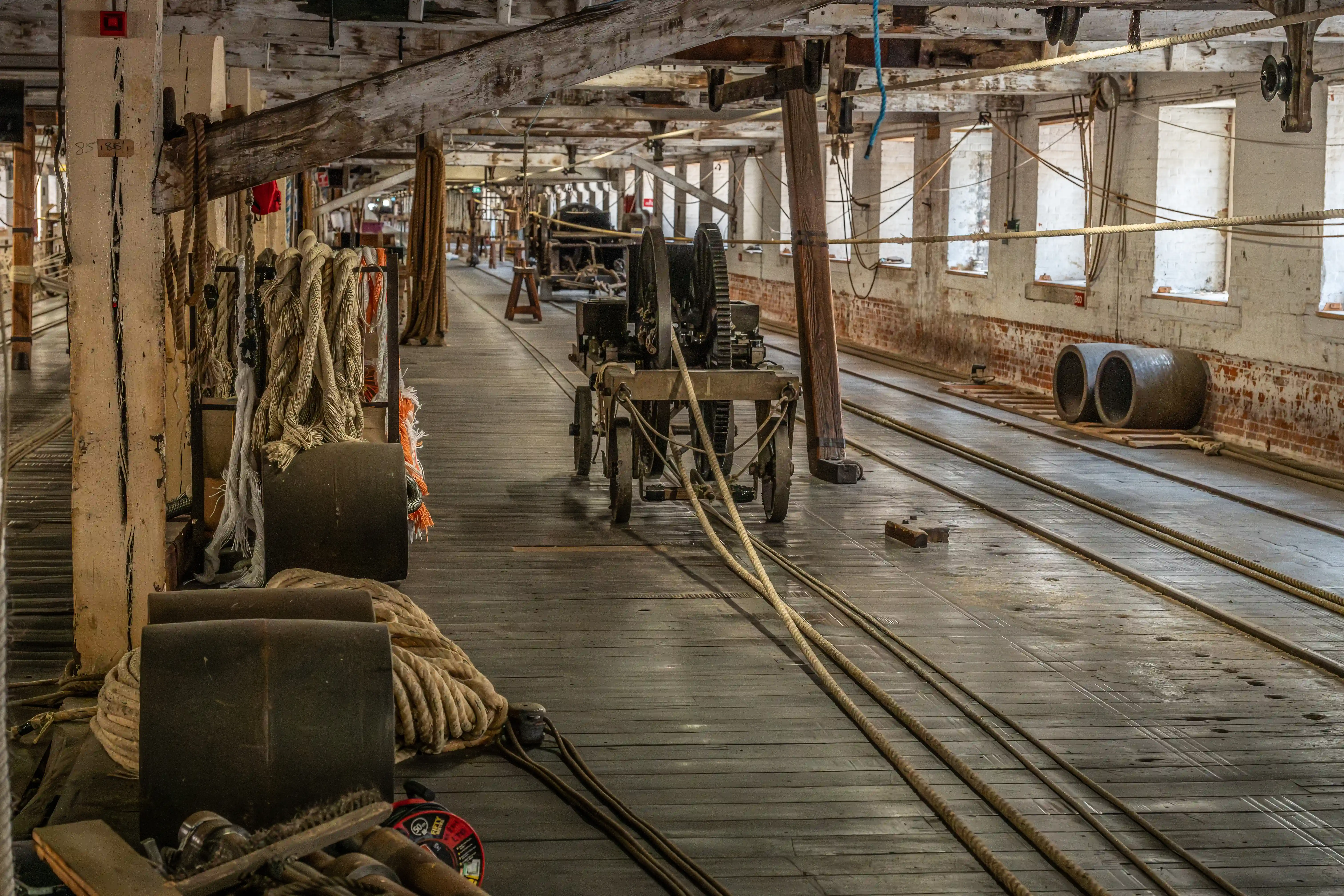 Interior view of an old rope walk with wooden floors, exposed beams, various coils and lengths of thick rope, and industrial machinery on wheels. Sunlight filters through large windows on the right.