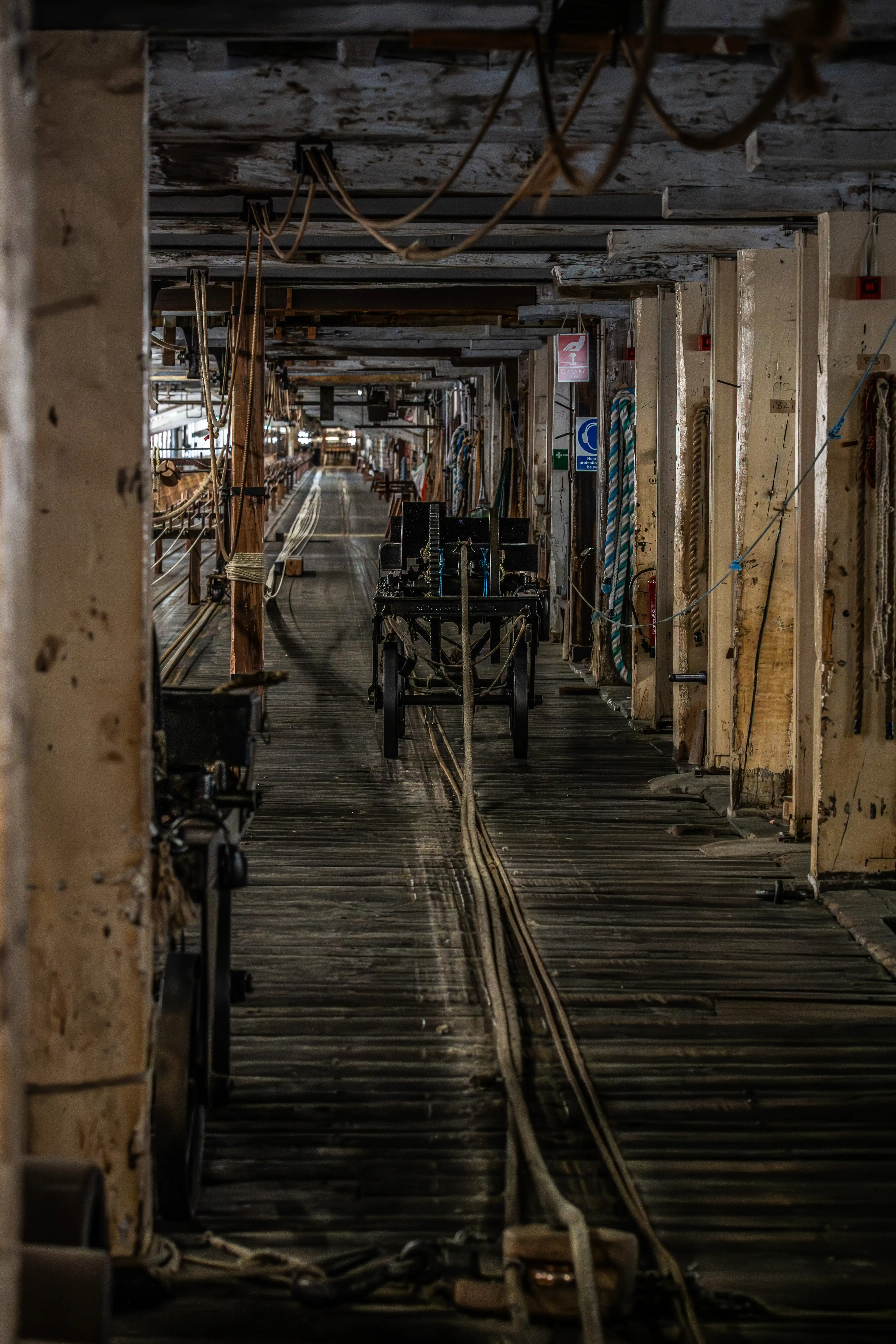 An old, empty factory interior with a wooden floor, tracks, and a wheeled cart, featuring hanging ropes and a sense of industrial abandonment.