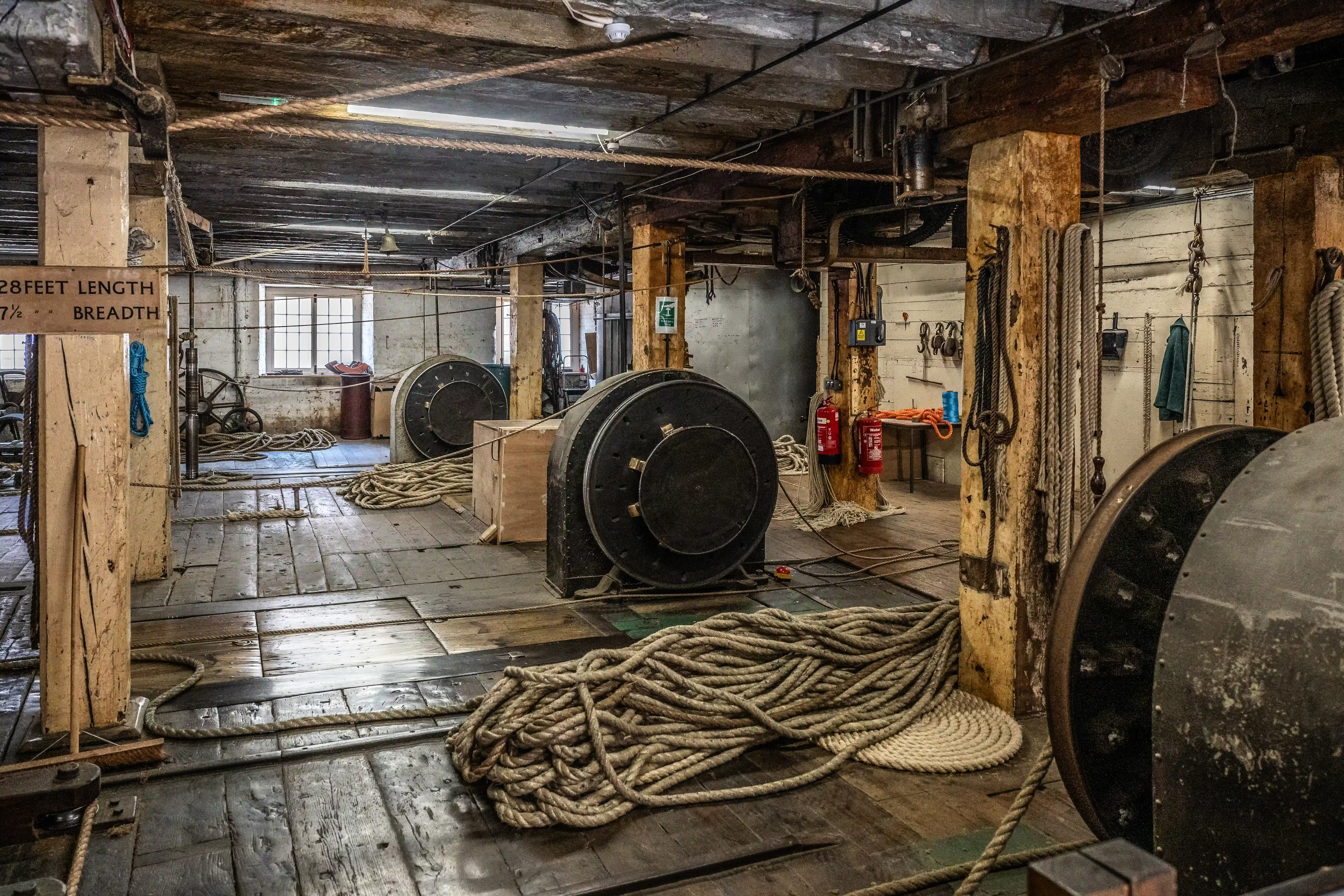 An interior view of an old industrial workshop, with large coiled ropes, winding machinery, wooden pillars, and a sign indicating rope measurements.