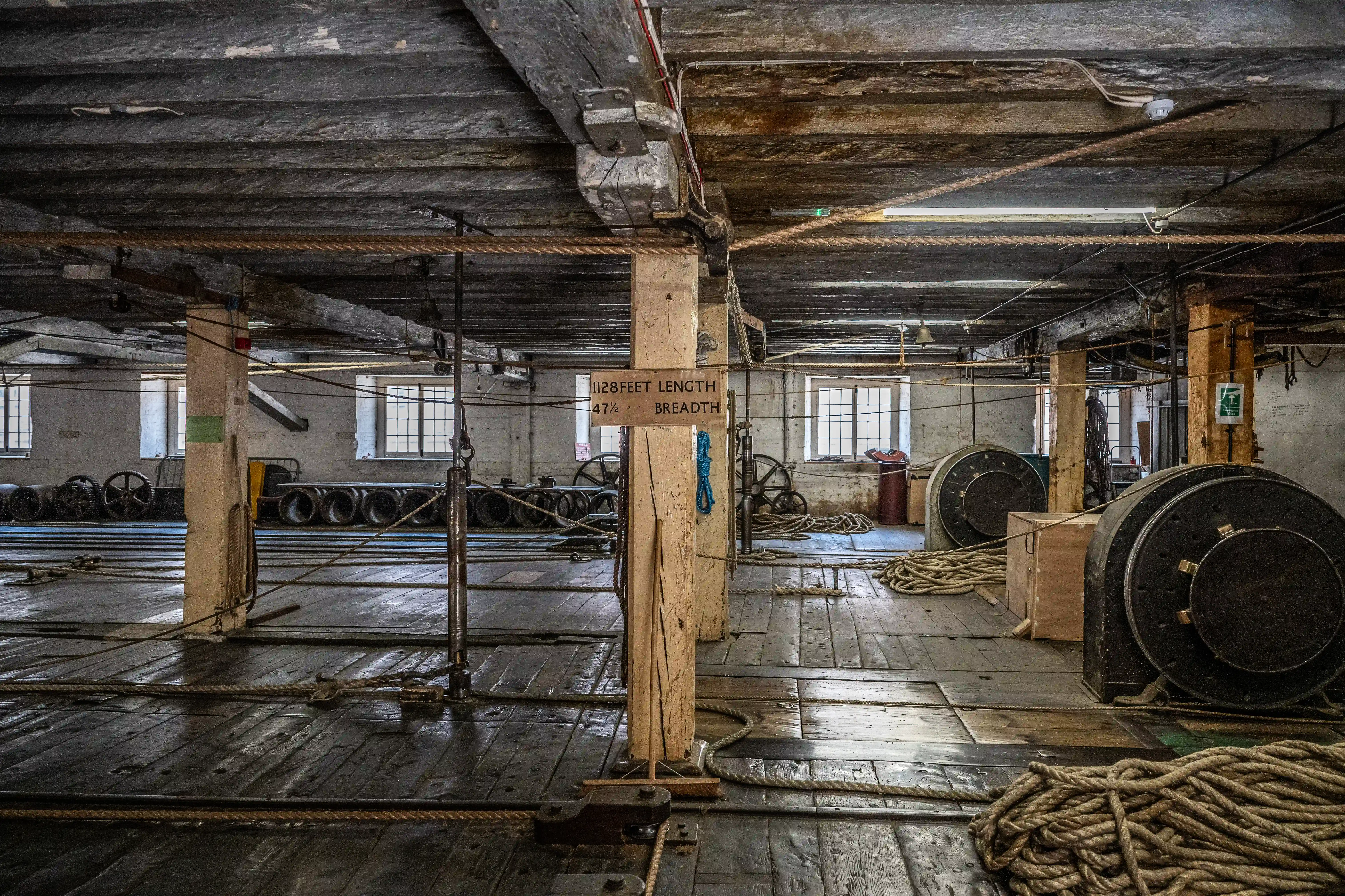 Interior of an old industrial building showing a wooden floor, exposed beams, large spools of rope, and windows.