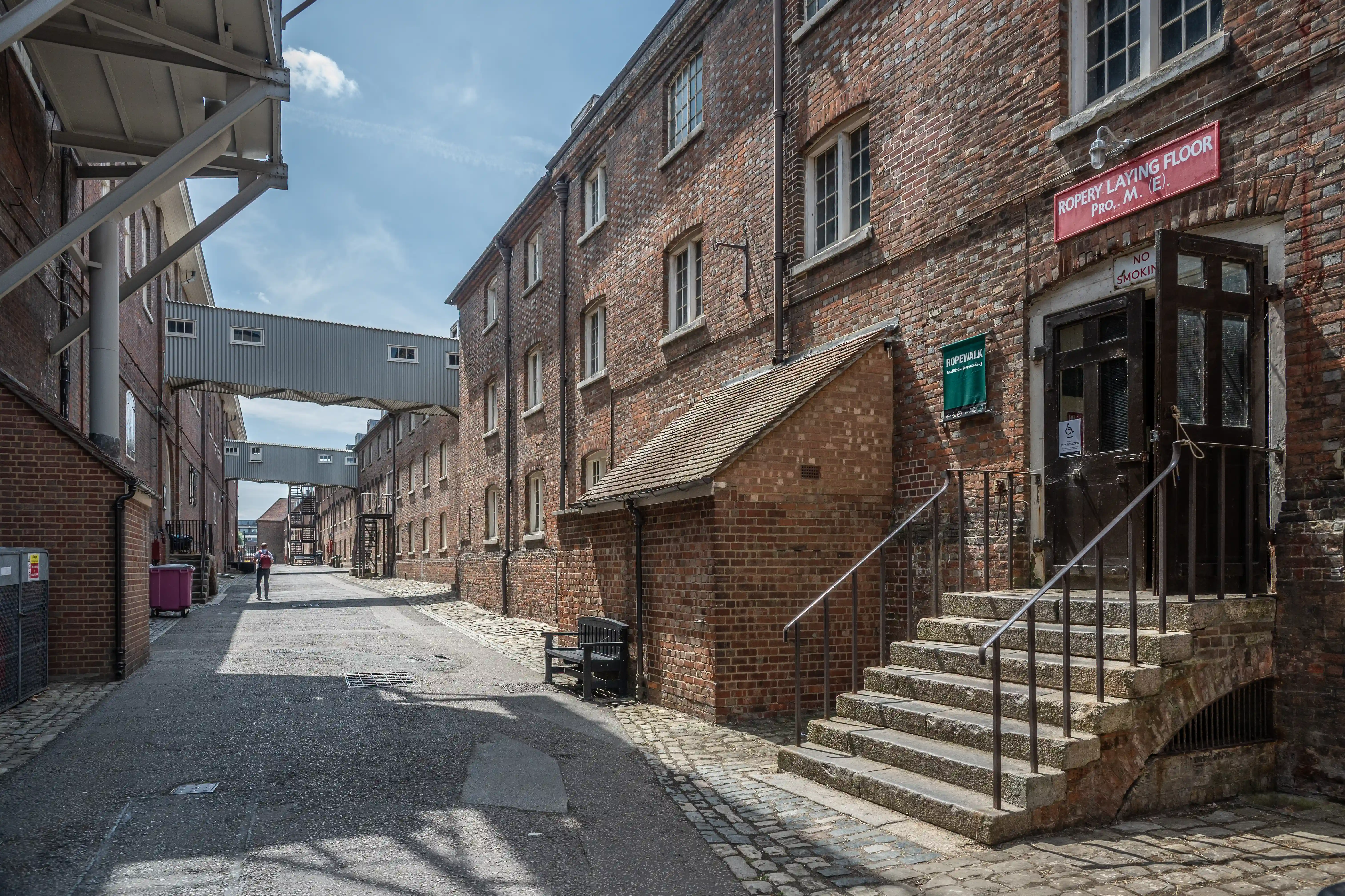 An exterior view of a historic industrial complex with red brick buildings connected by modern metal skywalks over a paved alleyway.