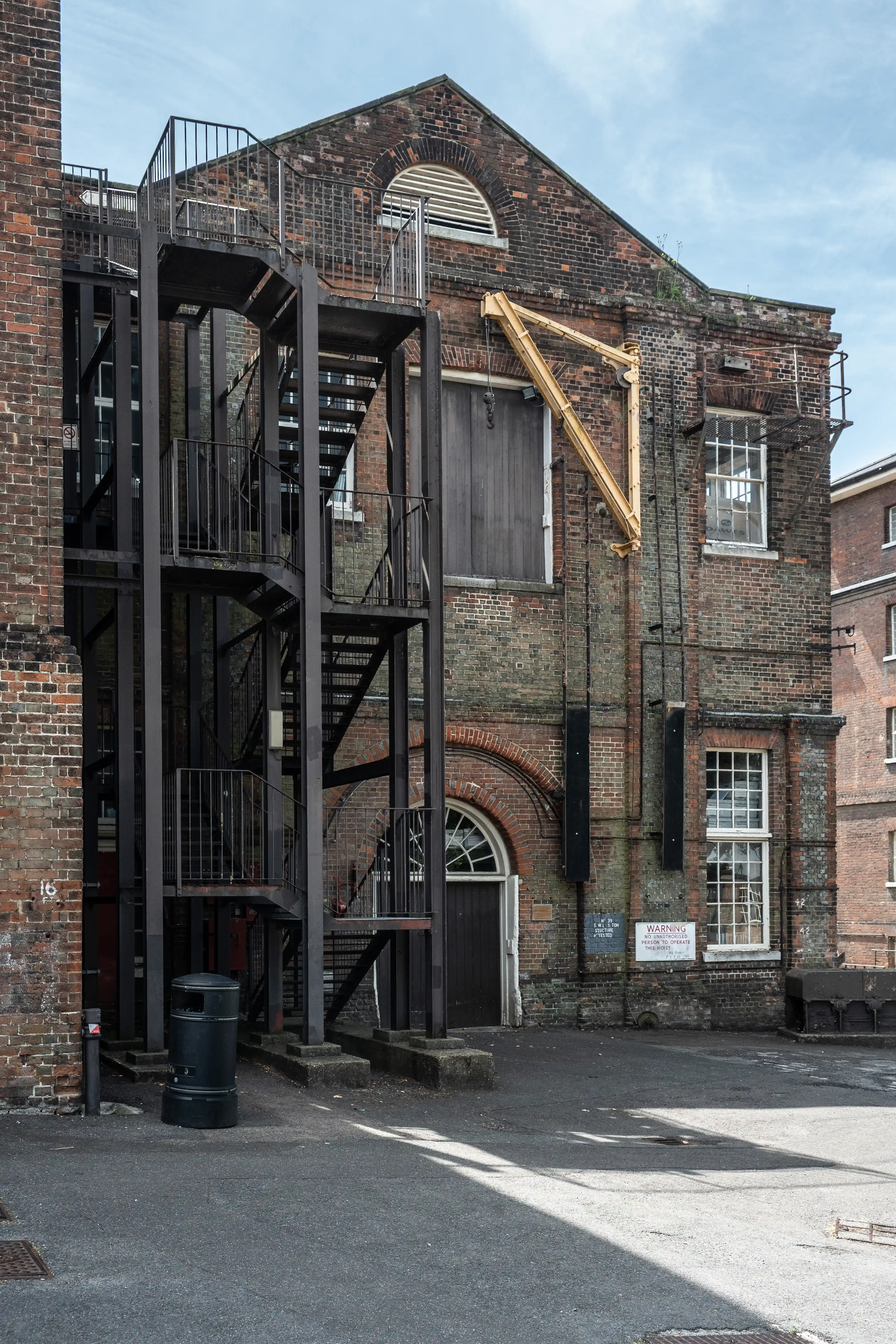 A multi-story red brick industrial building with a prominent black metal fire escape system and a yellow gantry crane attached to its side.