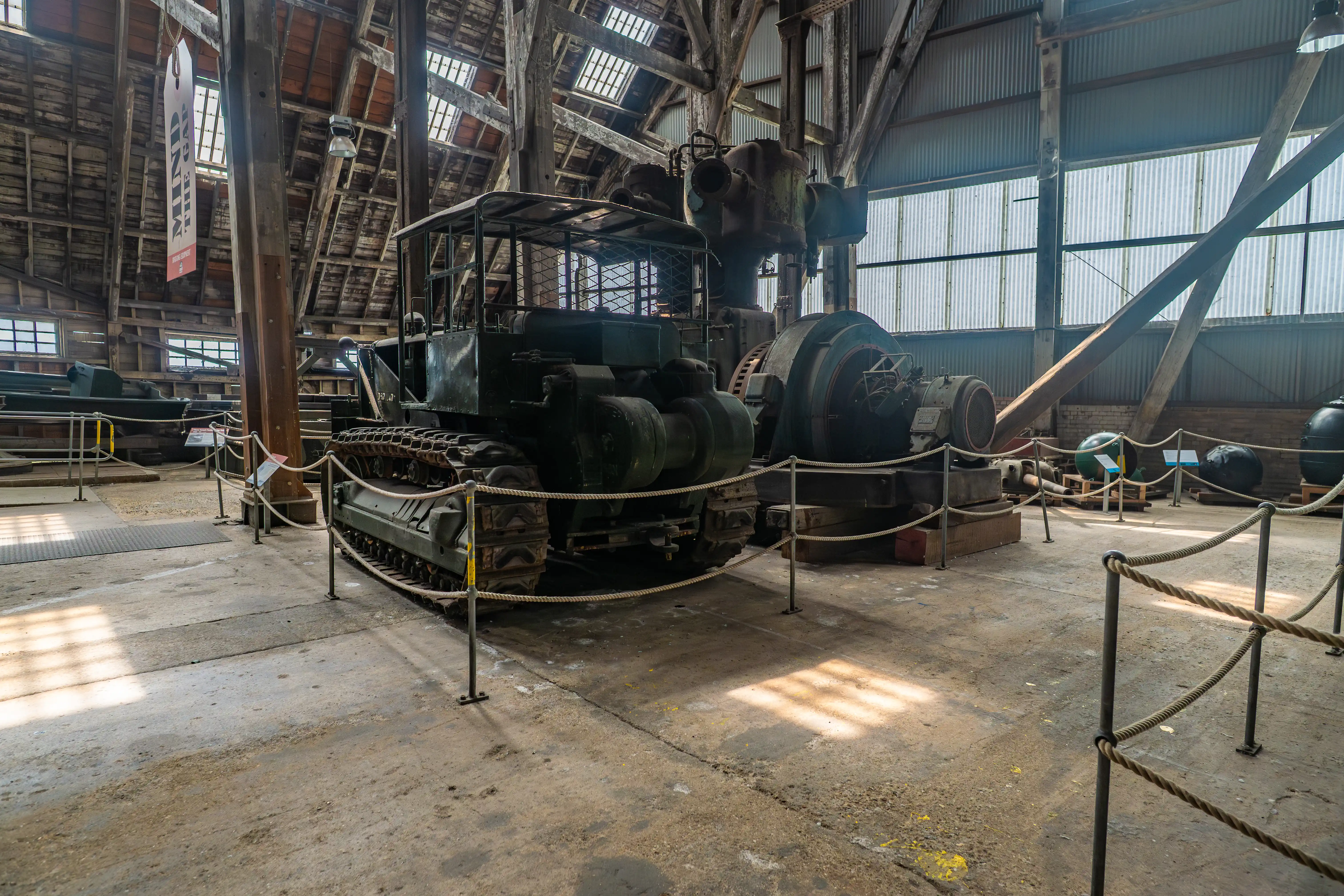 A large, green tracked vehicle and industrial machinery are on display in a dimly lit hangar with wooden beams and corrugated metal walls. Rope barriers are in the foreground, and sunlight streams through the windows.