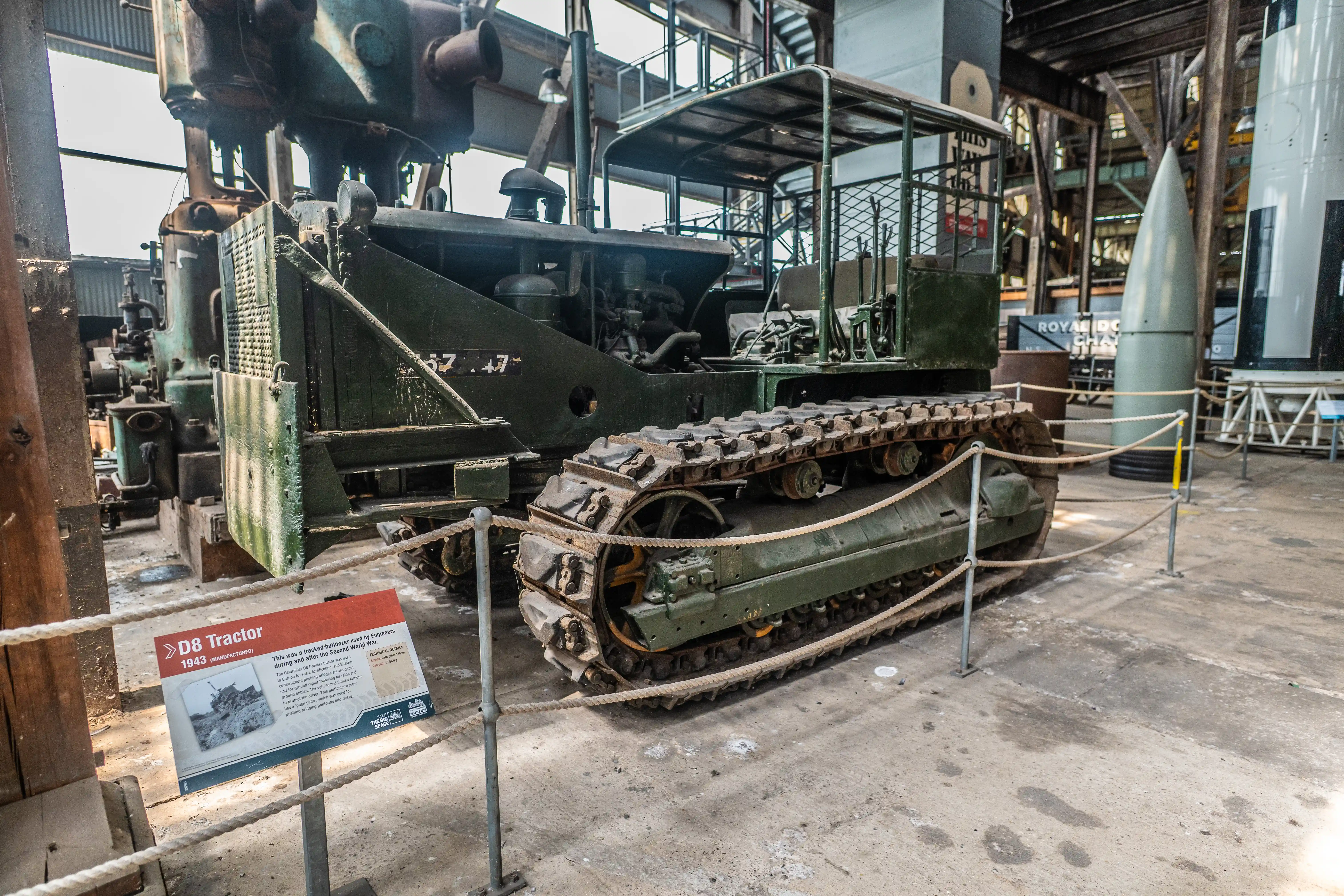 An olive-green, tracked D8 tractor from 1943 is displayed indoors. A descriptive sign is in the foreground. Other large industrial items and a rocket are visible in the background.