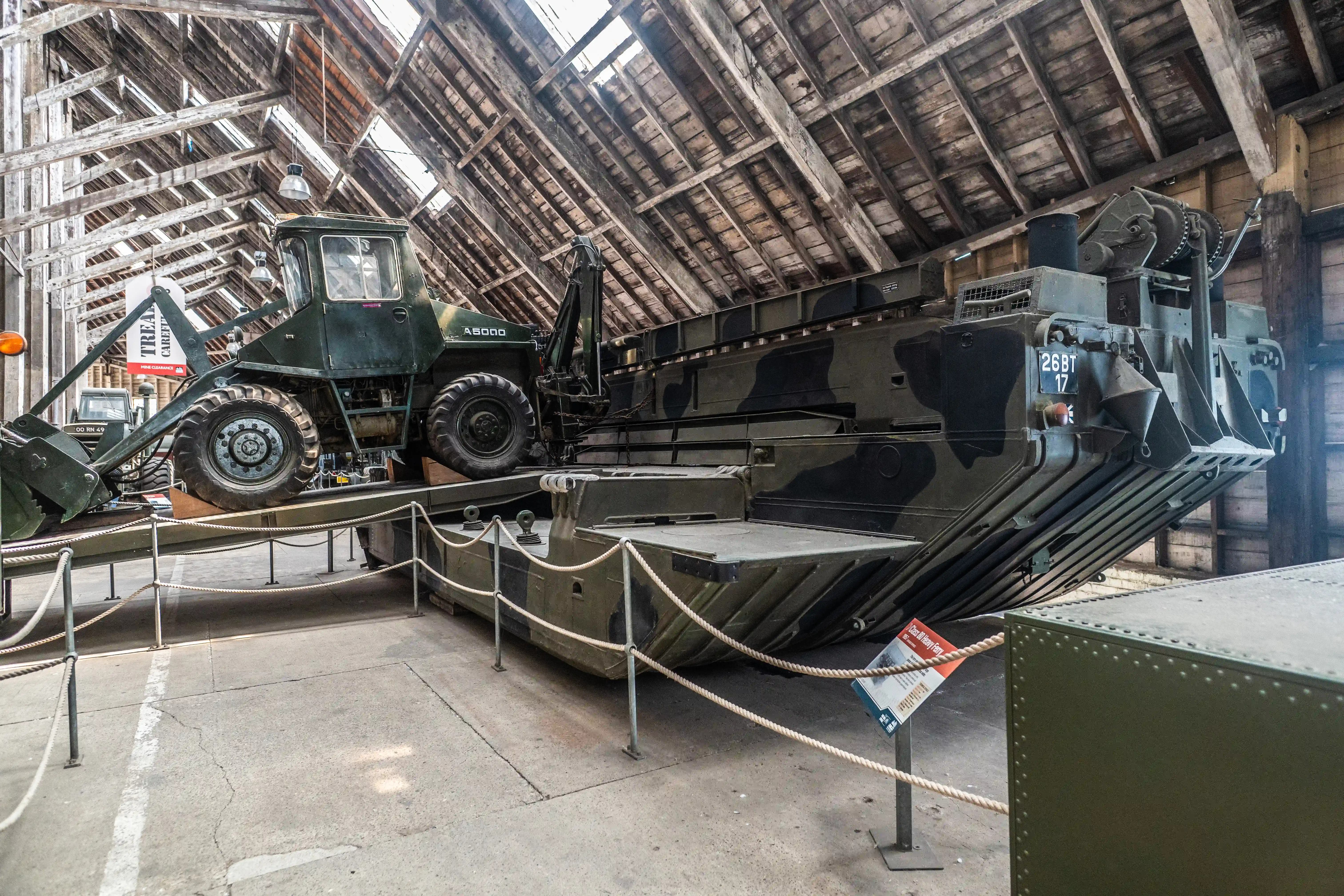 A dark green, camouflage-painted military amphibious vehicle, a Class 80 Heavy Ferry, rests on display in a wooden hangar. A wheeled loader vehicle is positioned on its deck. Rope barriers surround the exhibit.