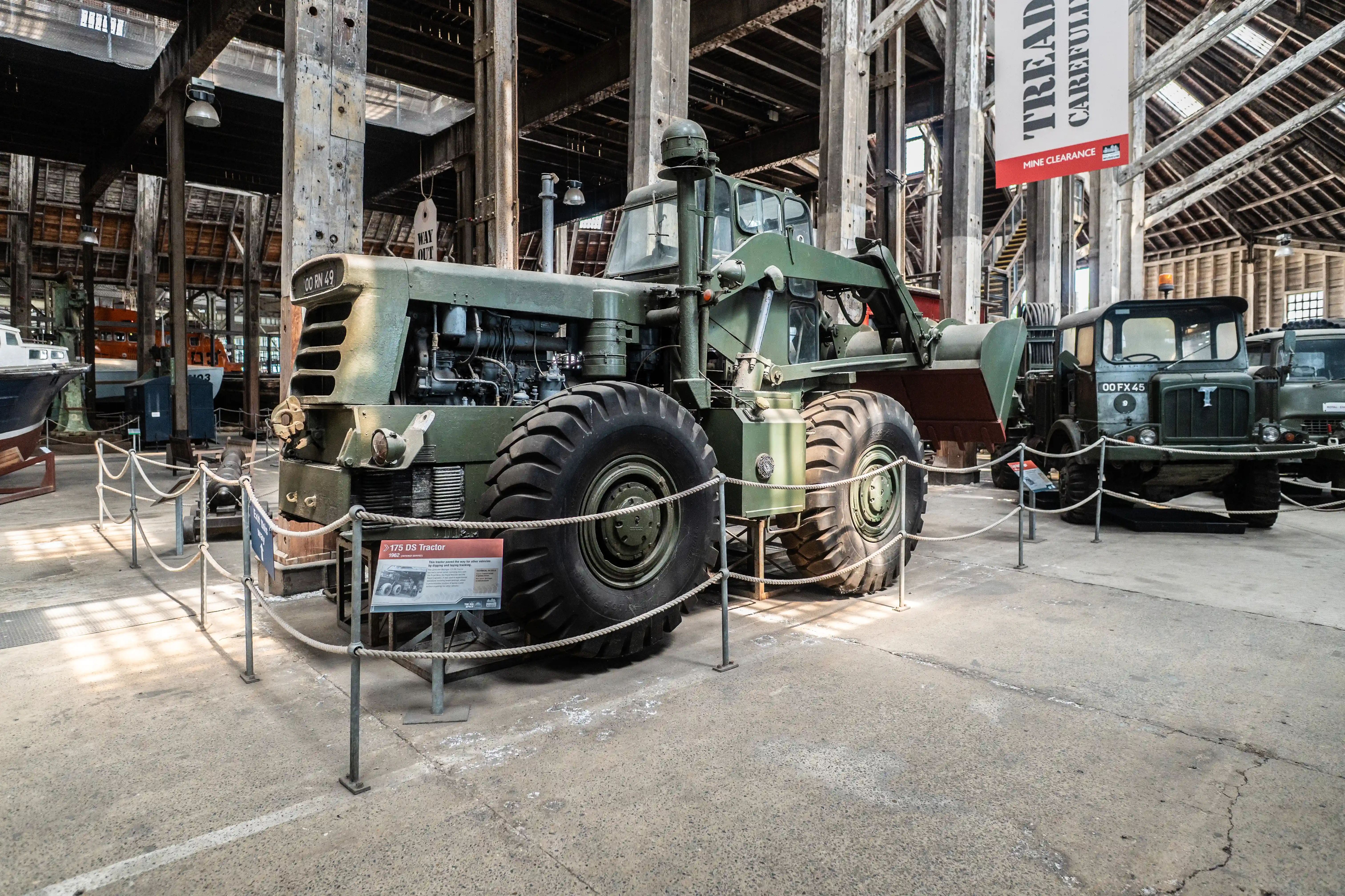 An olive-green military tractor with oversized tires and a front loader is displayed in a museum warehouse. Two military trucks are parked behind it. Ropes and stanchions guide visitors, and a banner reading TREAD CAREFULLY MINE CLEARANCE hangs overhead.