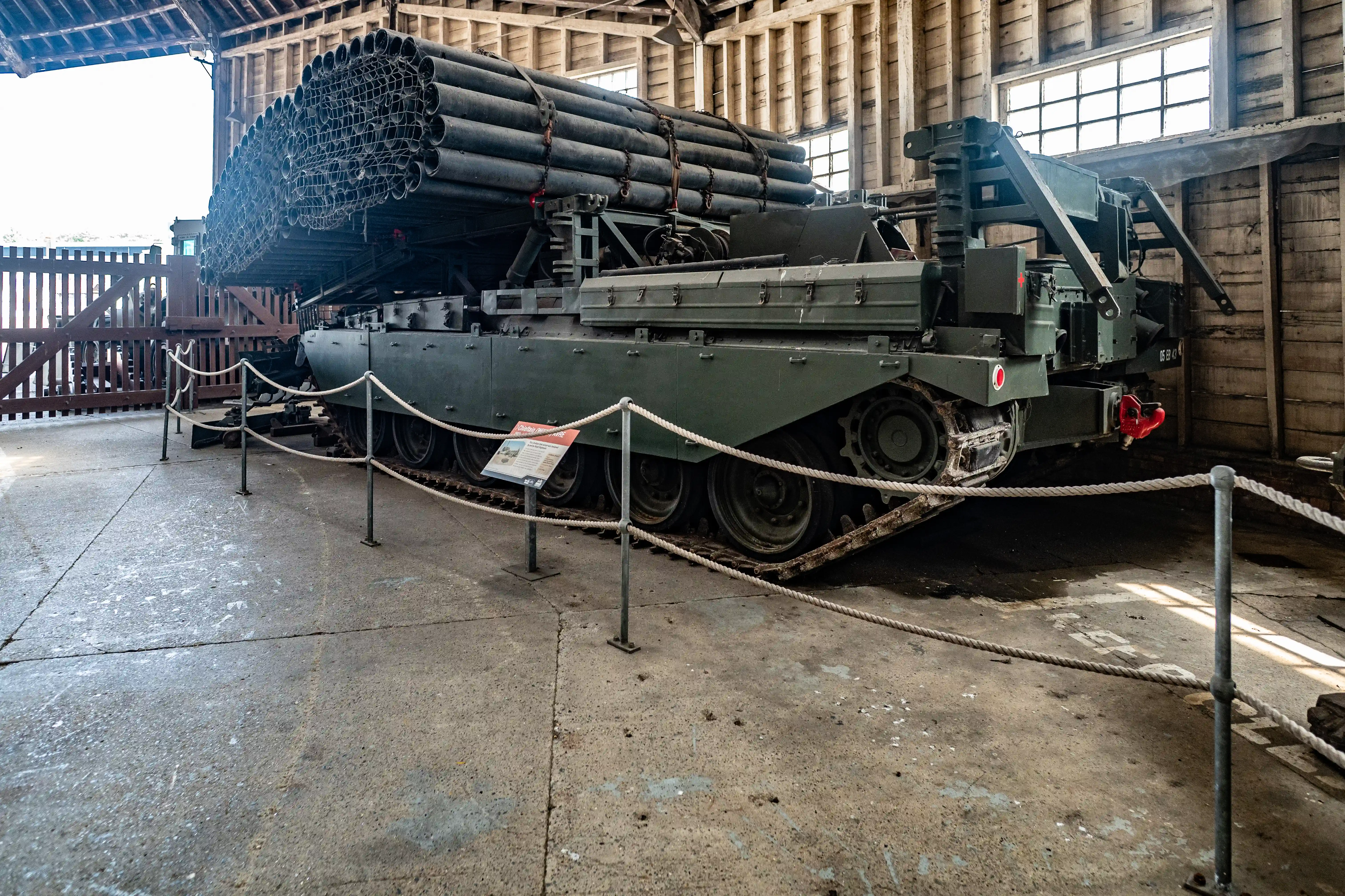 A tracked military vehicle with a large bundle of tubes mounted on top, displayed inside a rustic wooden building.