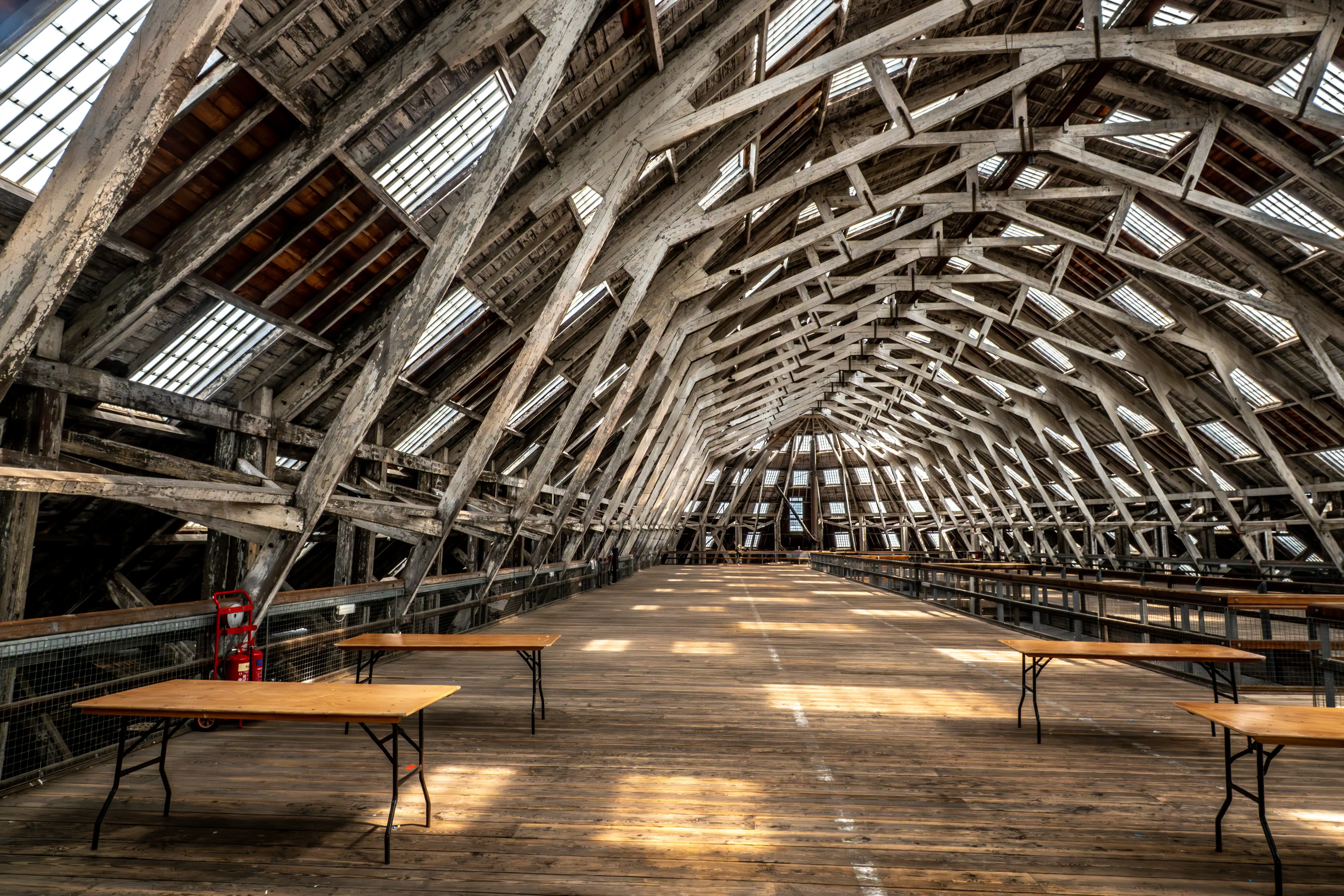 Interior view of a large, aged wooden arched roof structure with numerous skylights, a wooden floor, and several empty folding tables.