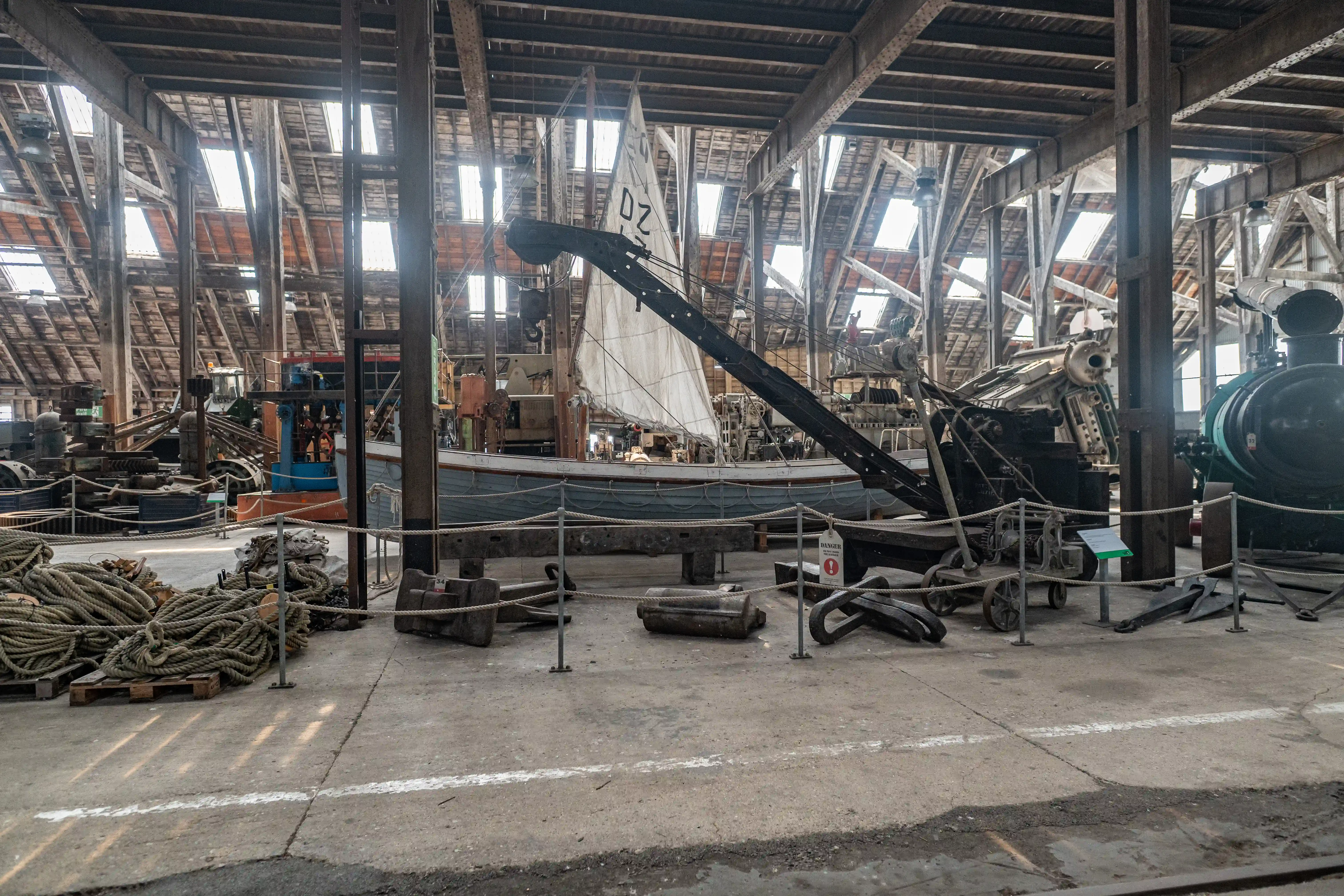 A museum exhibit displaying a boat with sails, a large crane, and various industrial maritime artefacts inside a large hall with a wooden beamed roof.