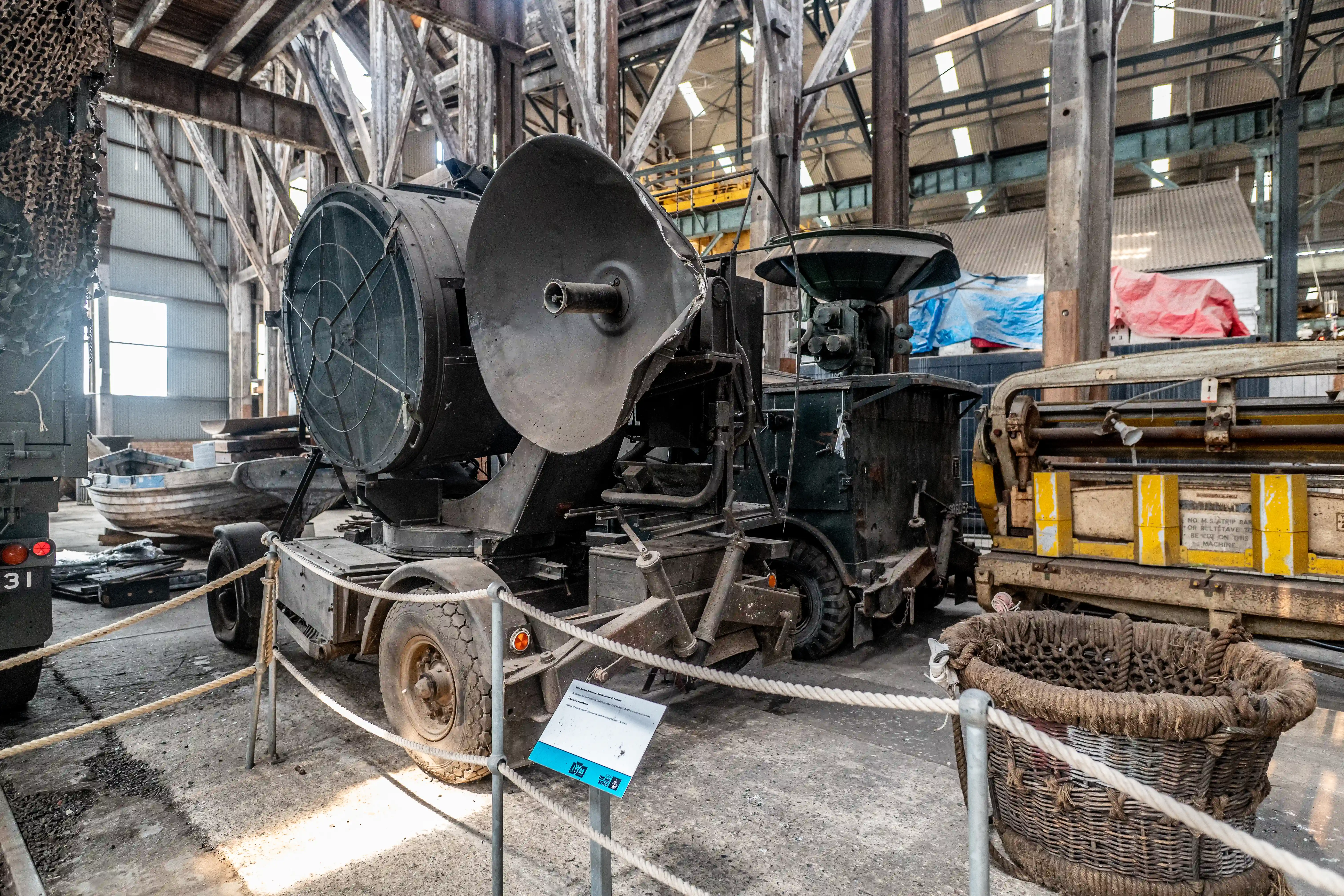 A vintage military searchlight with a large dish sits on a trailer inside a large, wooden-beamed warehouse.
