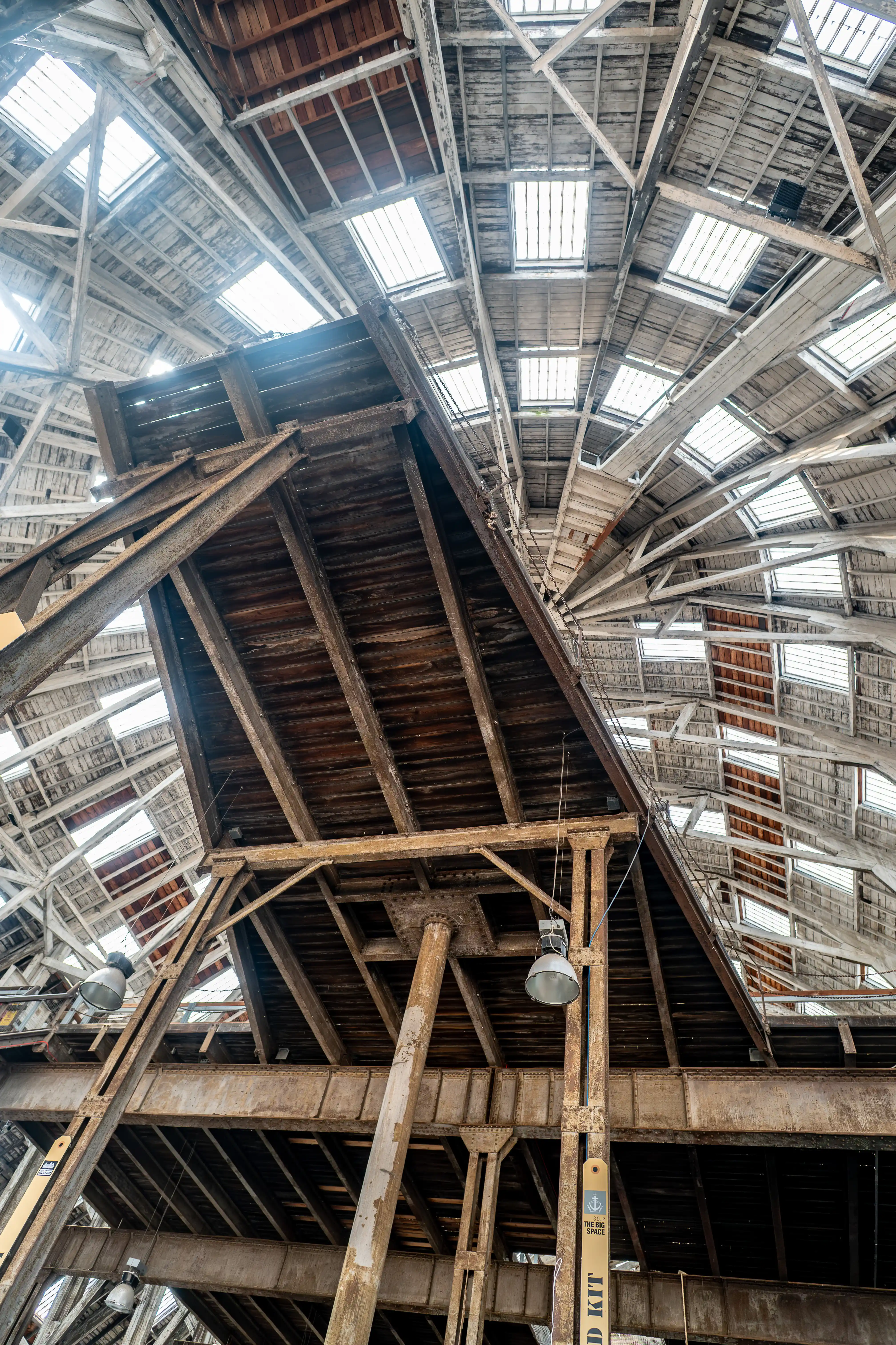 View from below inside a large industrial structure showing a geometric pattern of wooden rafters and steel beams against a backdrop of many rectangular skylights.